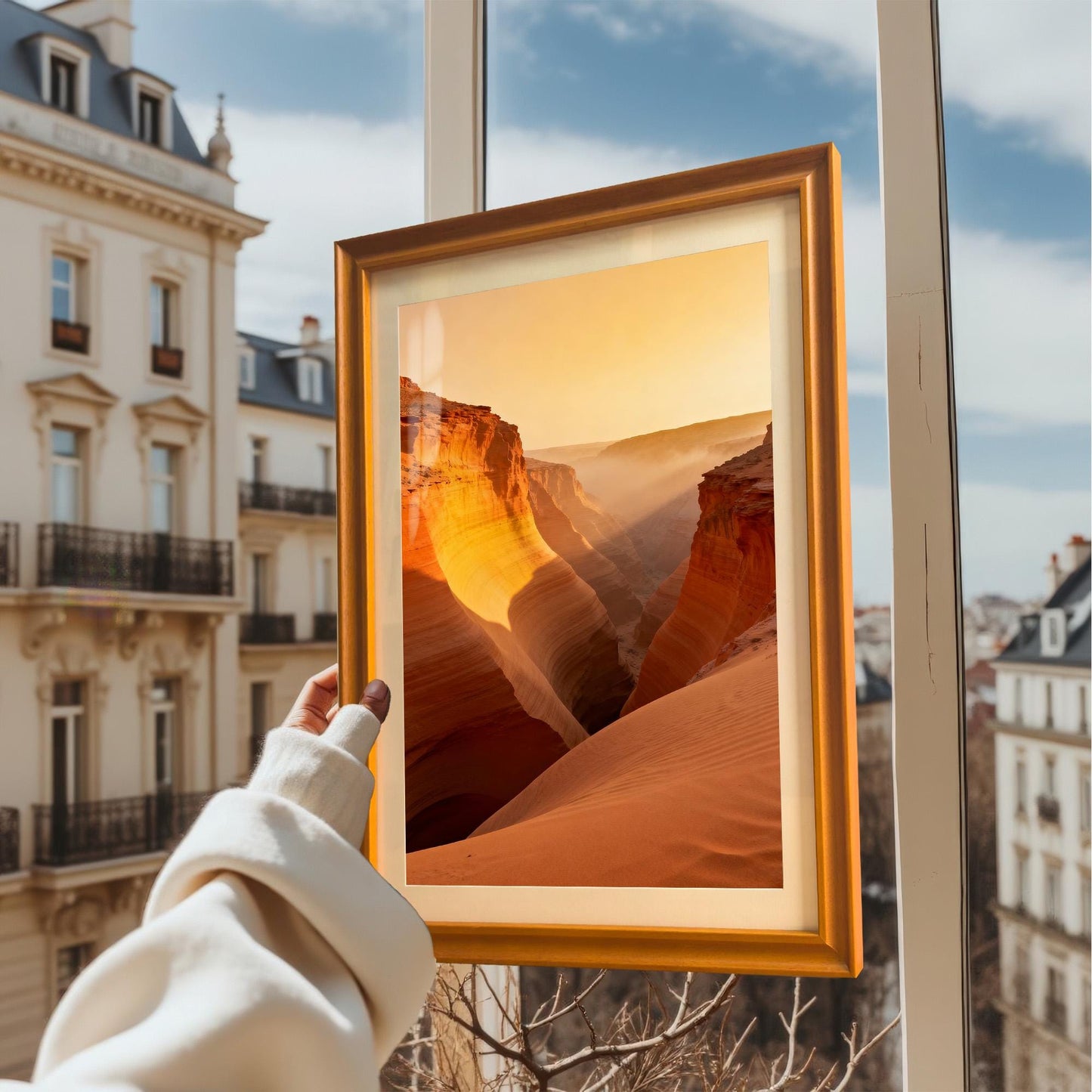A hand in a cream sweater holds up a framed art print of the canyon sunset against an exterior window view of a European city. The frame is Orange-colored wood.