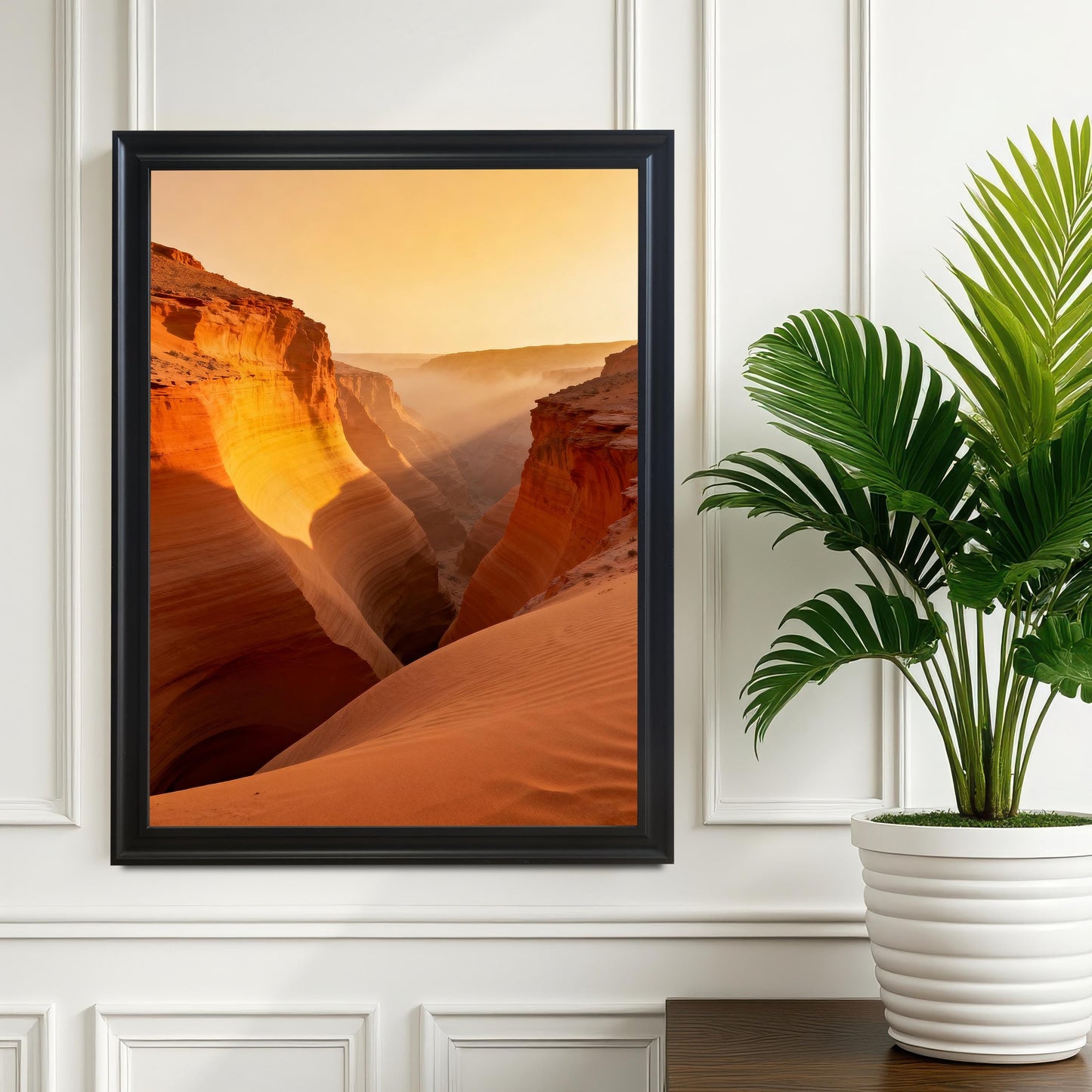 Framed print of the vibrant canyon with mist and sunlight hanging on a white paneled wall above a dark wooden surface, next to a potted palm plant. The frame is Black wood.