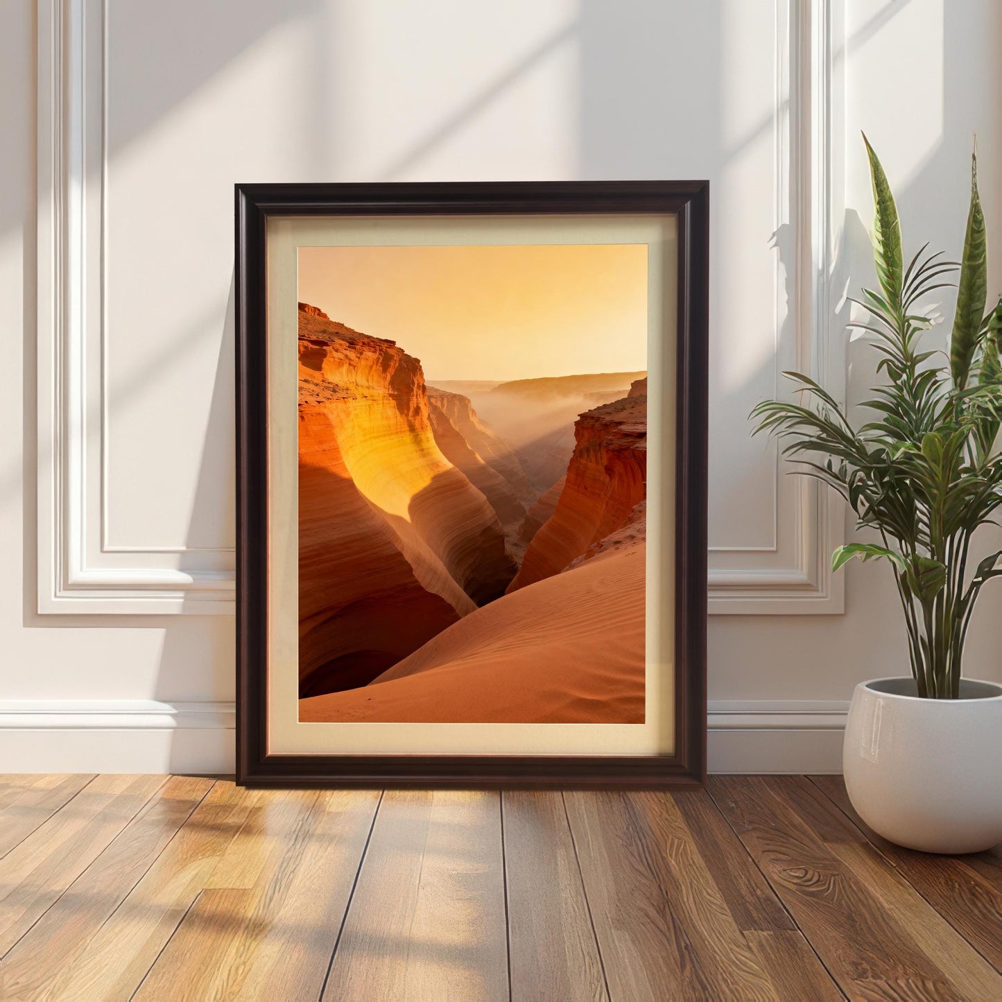 Framed print of a slot canyon landscape at sunset showing layered orange rock and sand, leaning against a white paneled wall on a wood floor next to a snake plant. The frame is Coffee-colored wood with matting.