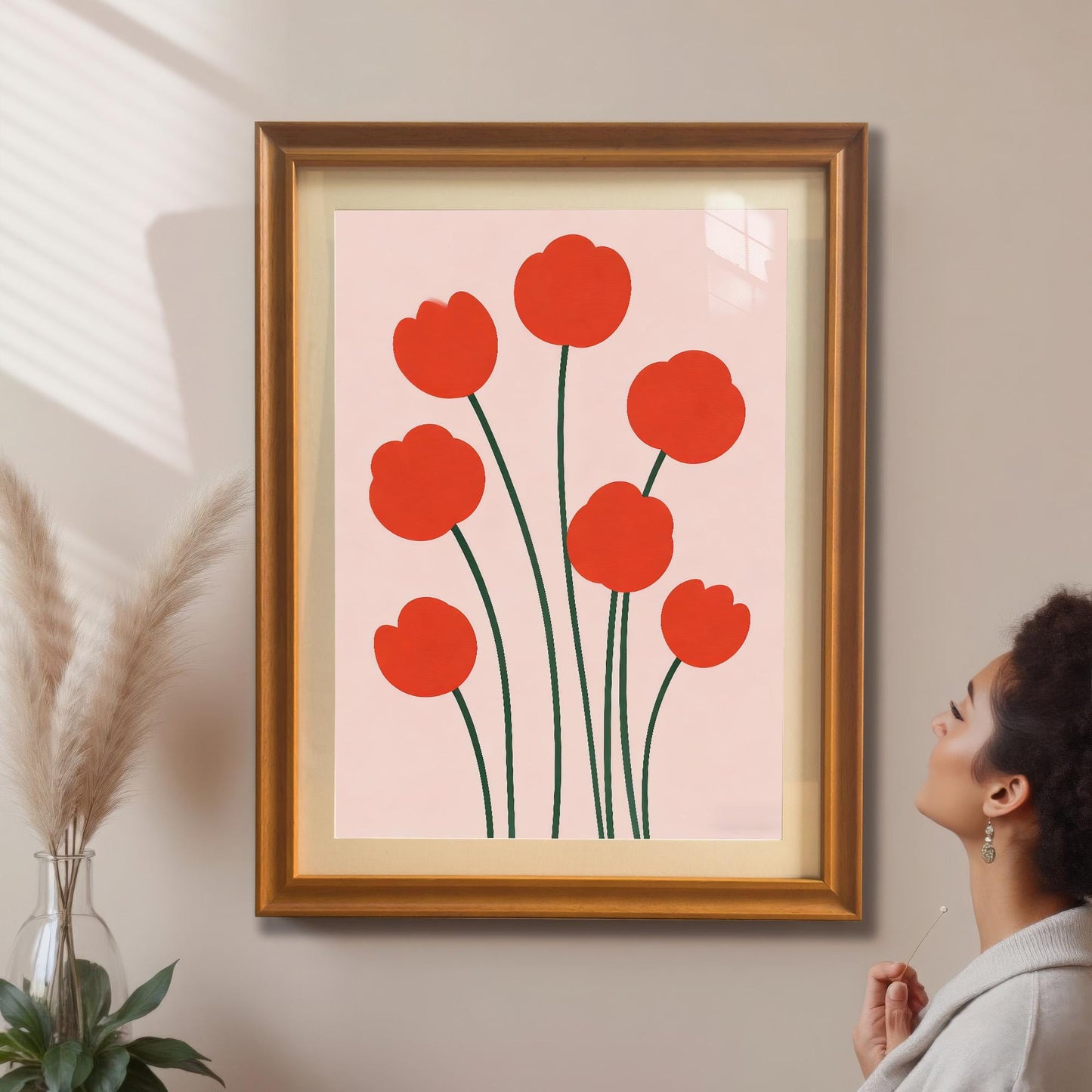 A woman admires a framed print of seven minimalist red flowers hanging on a tan wall. The frame is Orange-colored wood. Pampas grass is visible in a vase to the left.
