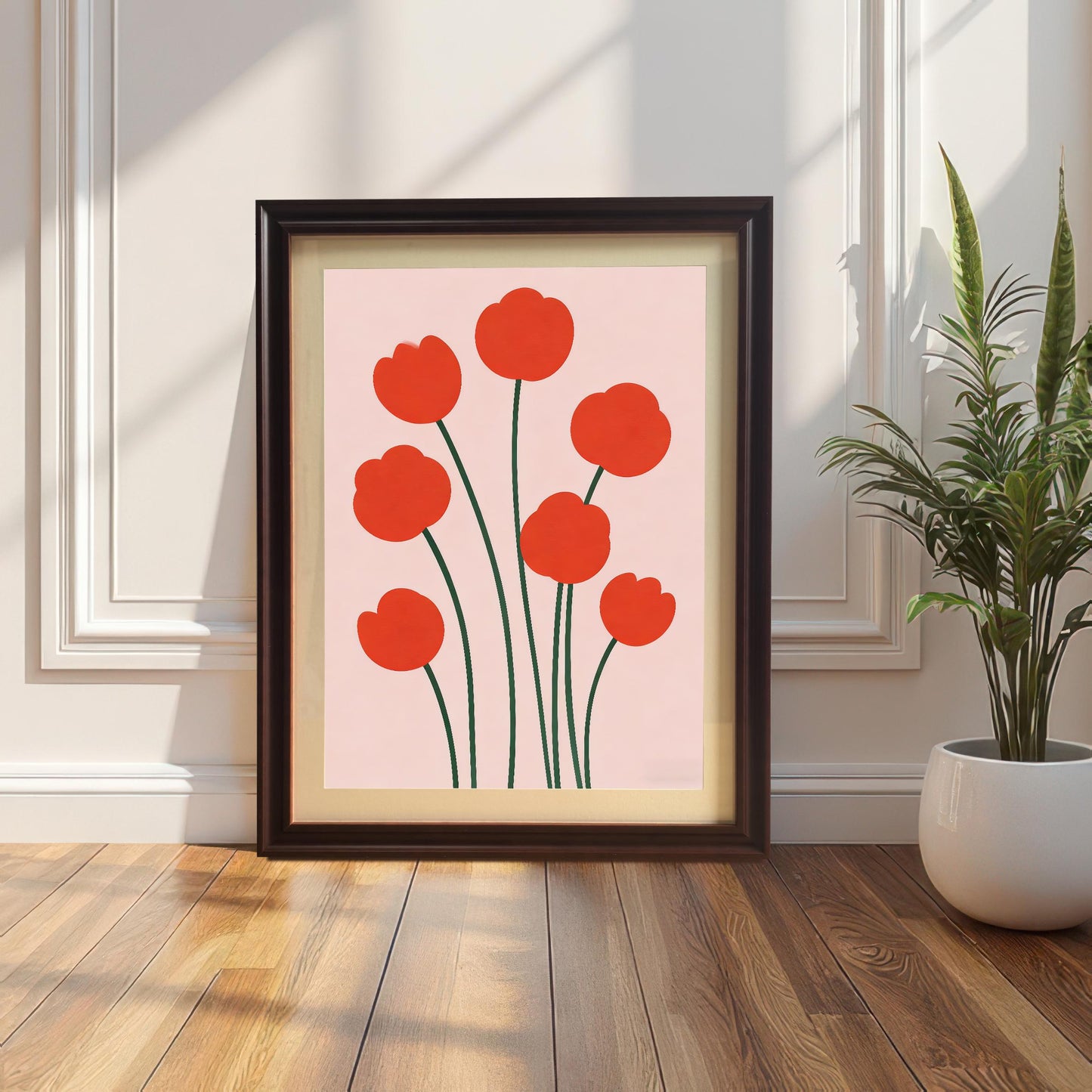 Framed print of seven red, minimalist flowers on a pale pink background leaning against a white paneled wall on a wood floor next to a snake plant. The frame is dark Coffee-colored wood with matting.
