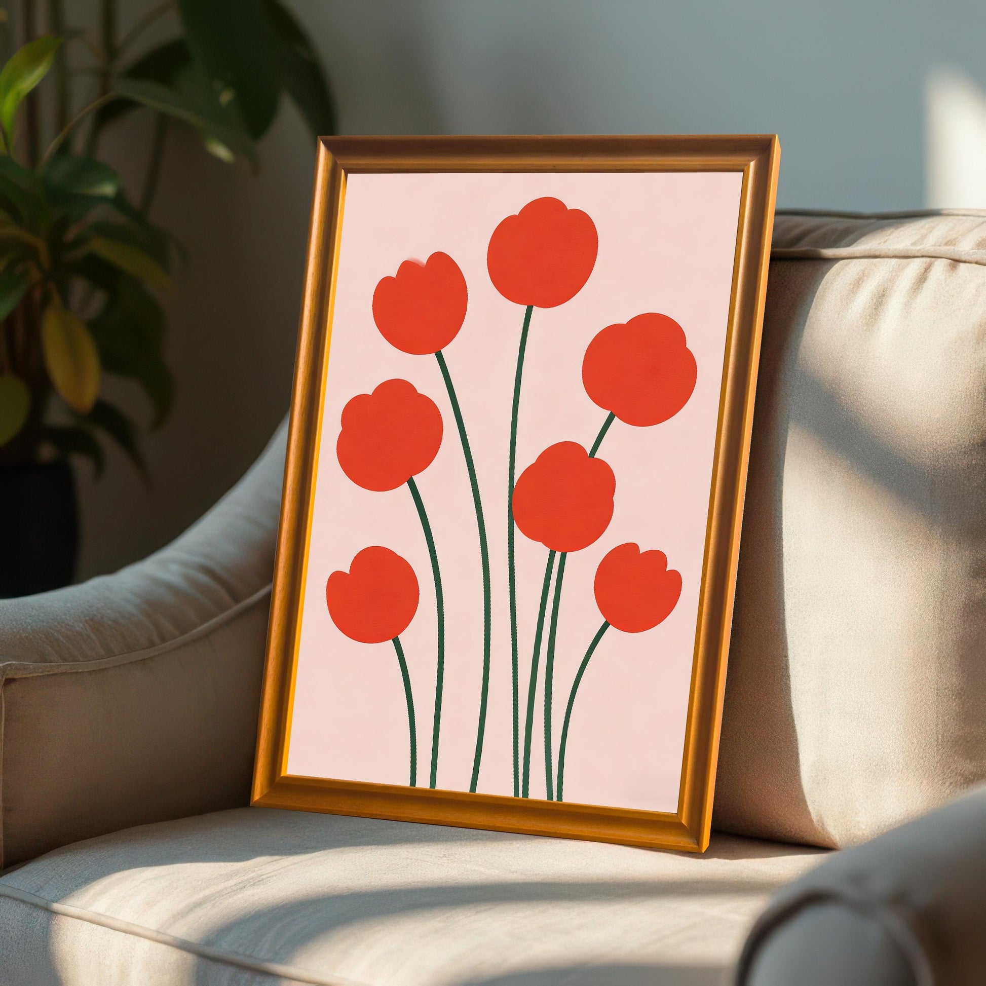 Framed print of red poppy-like flowers in an Orange-colored wood frame leaning on a plush, light-colored armchair bathed in sunlight.
