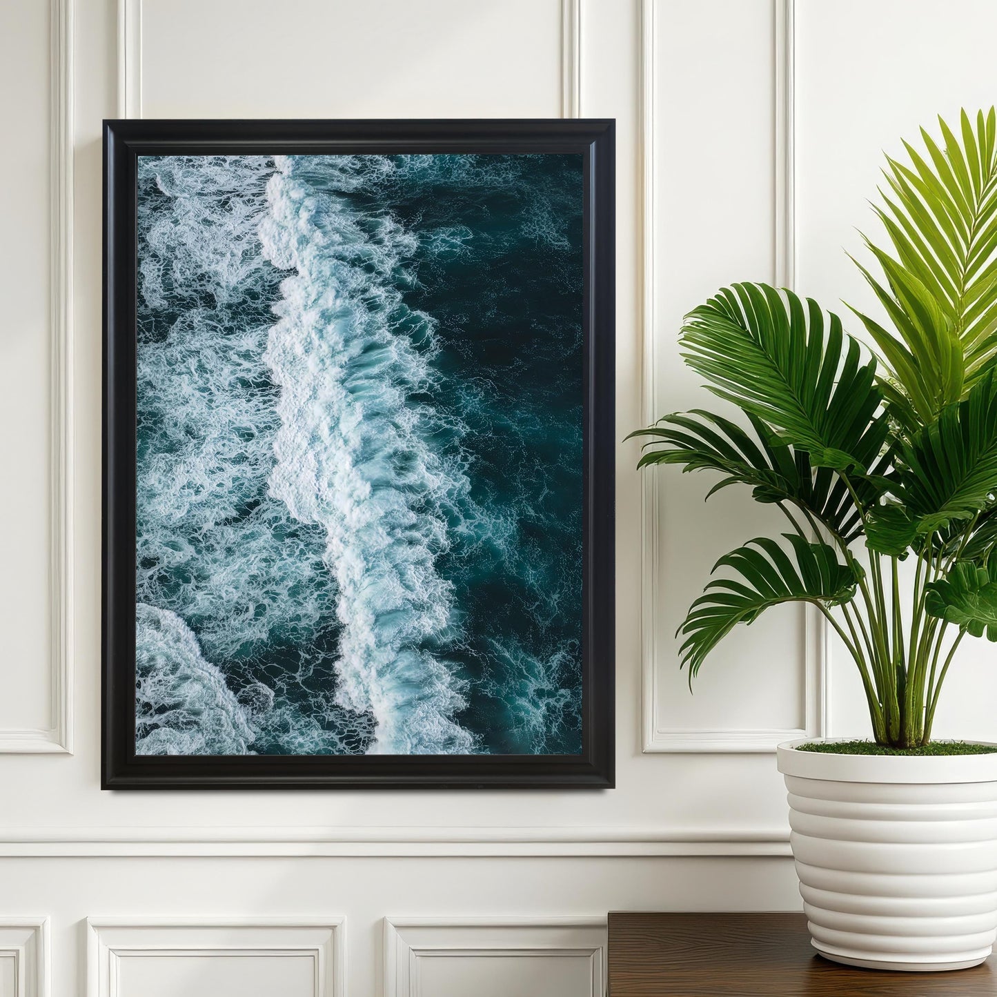 Large black framed print of a dark blue ocean wave with white foam, hanging on a white wall next to a potted green plant for a modern decor aesthetic.