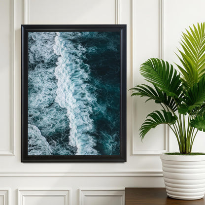 Large black framed print of a dark blue ocean wave with white foam, hanging on a white wall next to a potted green plant for a modern decor aesthetic.