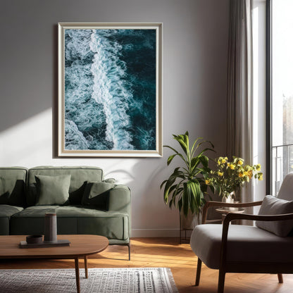 Large, portrait-oriented framed print of an ocean wave with a White/Silver wood frame (no mount), hanging above a green sofa in a modern, sunlit living room with a potted plant and natural wood furniture.