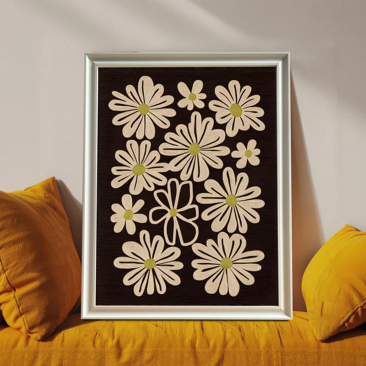 White framed art print of retro daisy flowers on a dark background, leaning against a wall between two yellow cushions.