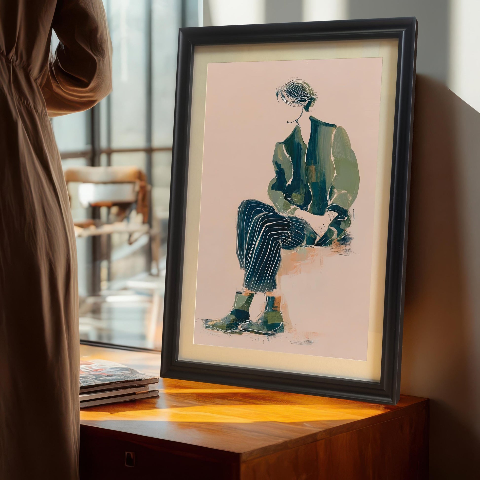 Lifestyle photo of the art print in a Black wood frame with a mat/mount, standing on a wooden cabinet near a large window, next to a person in a brown dress. The artwork shows a seated figure in green and dark striped clothing.