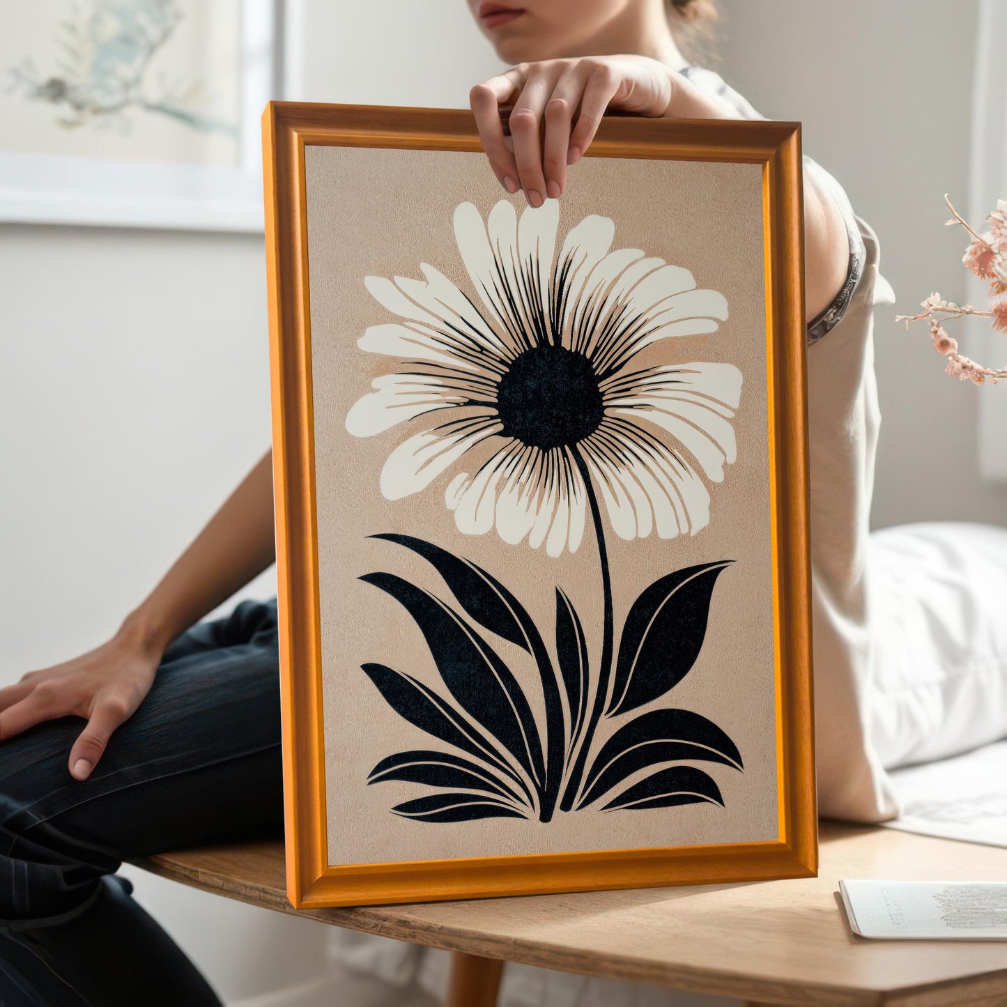 Lifestyle photo where a person is holding the framed print, showing the Orange wood frame. The artwork is a minimalist illustration of a white flower with a black center and black leaves on a beige background.
