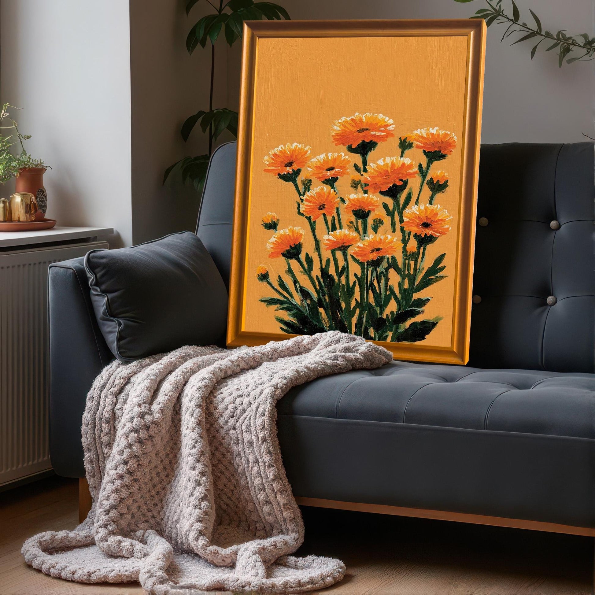 A framed orange floral art print in an Orange wood frame leaning against a dark gray tufted sofa with a knit beige throw blanket.