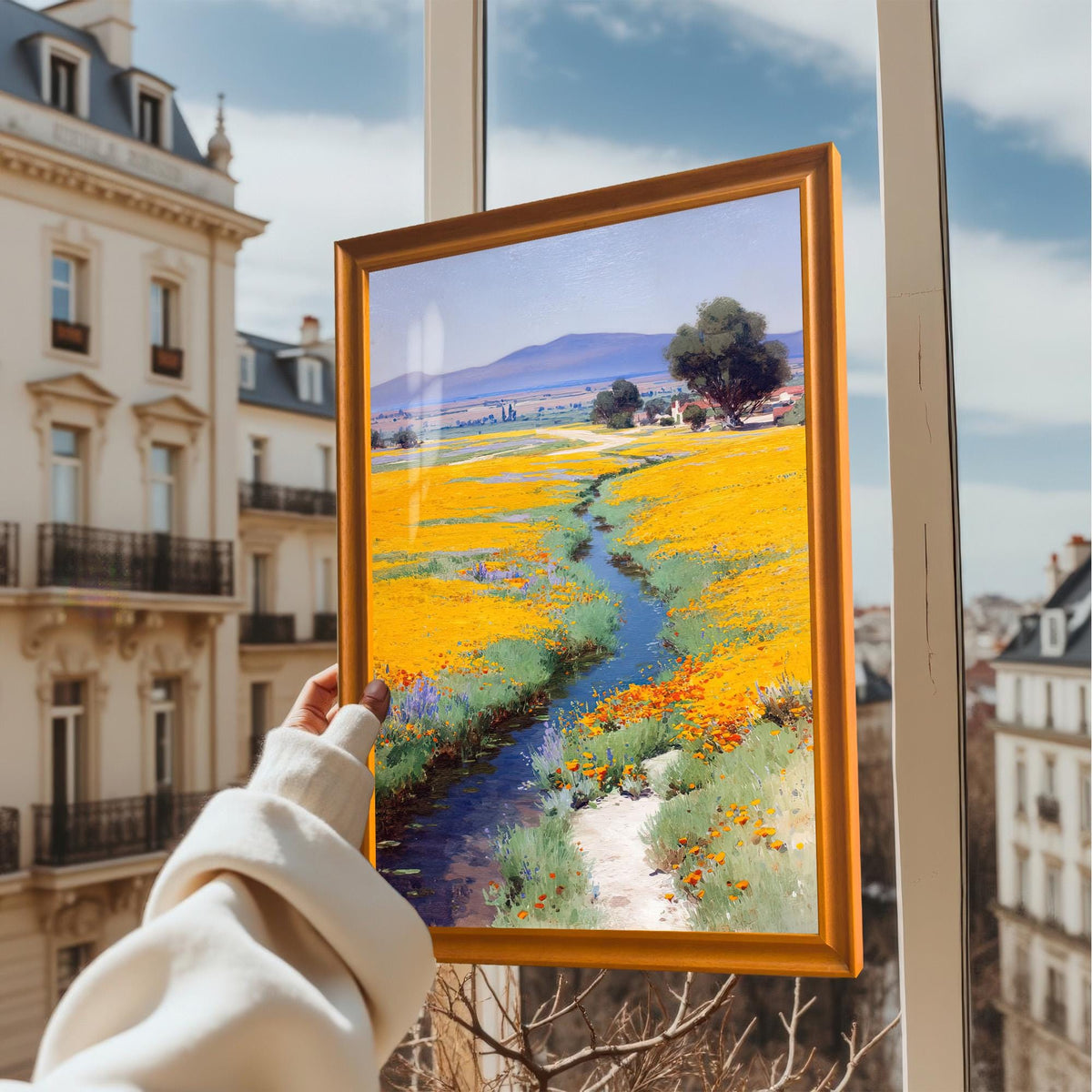 A hand holding up the framed art print (Orange Frame, No Mount) in front of a window overlooking a classic European city street scene.