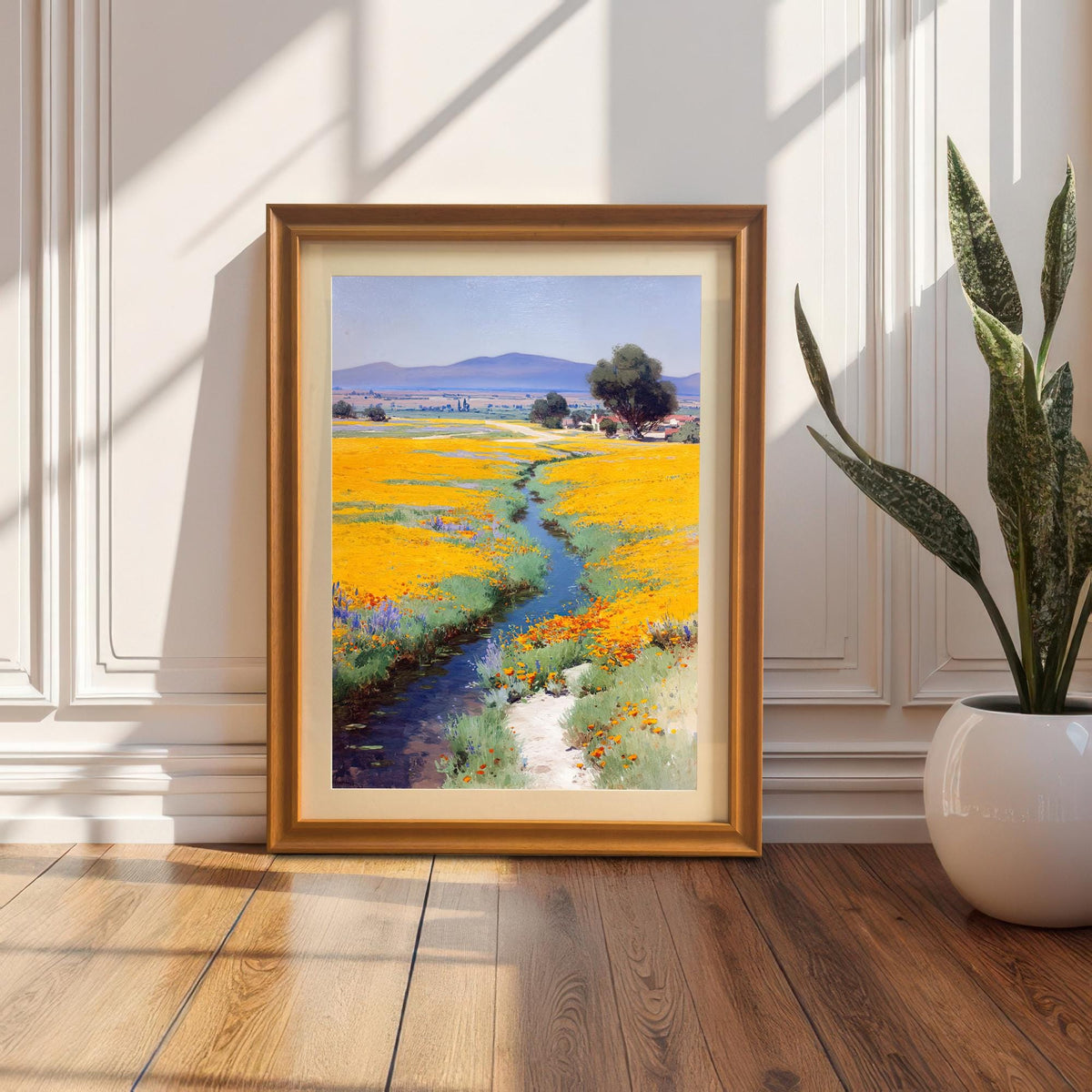 Framed art print (Orange Frame with Mount) leaning on a wood floor against a decorative white paneled wall next to a large potted snake plant, showing sun shadows.