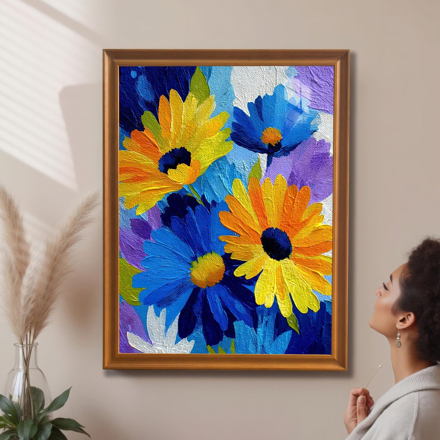 A woman looking up at the framed abstract blue and yellow flower print in an orange/gold frame with a mat/mount hanging on a beige wall, next to pampas grass decor.