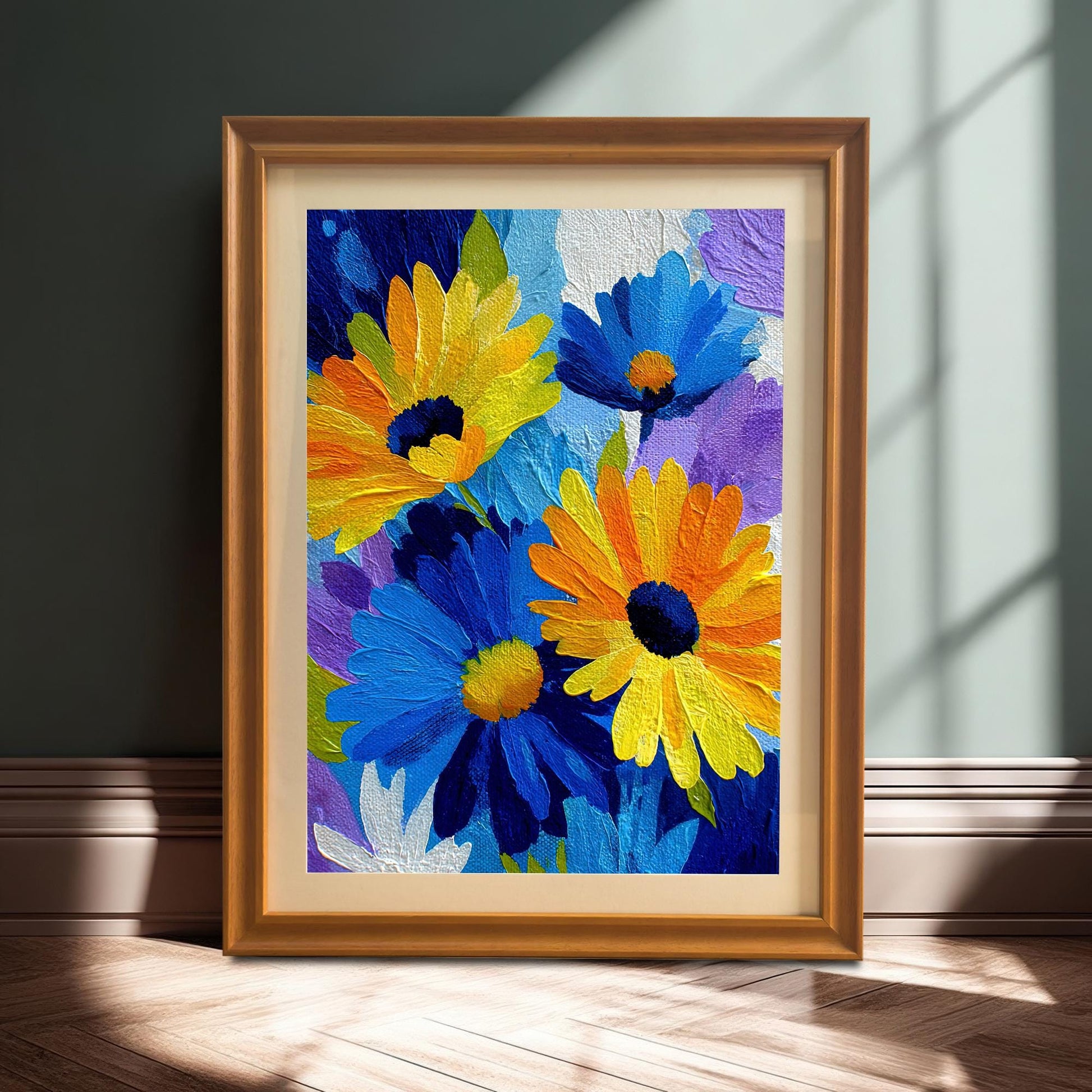 Framed print of an abstract blue and yellow flower painting with thick texture, shown in an orange/gold frame with a mat/mount leaning on the floor against a dark wall with sunlight and shadows.