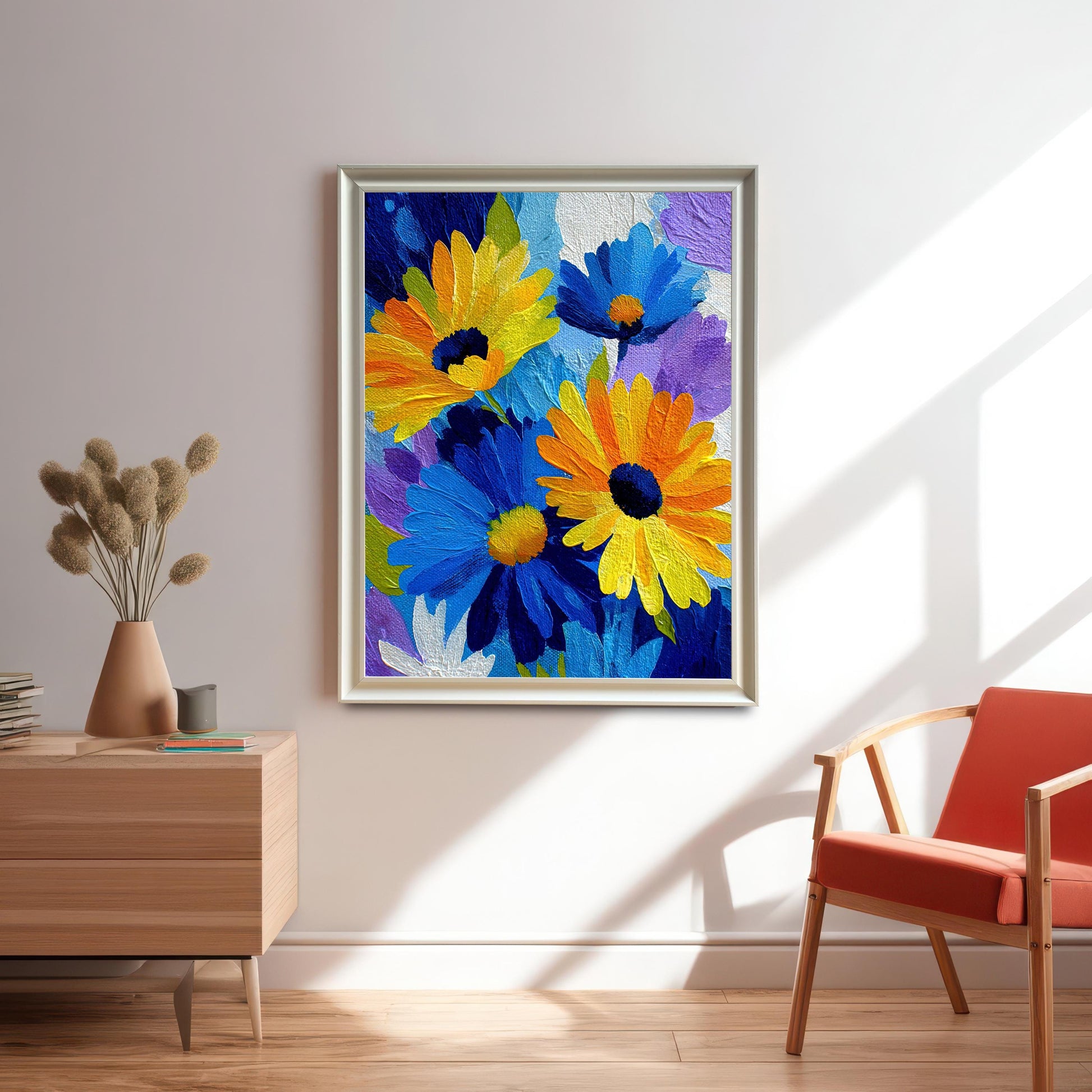 Large framed print of the vibrant blue, yellow, and orange floral artwork in a silver/light gold frame, hanging on a white wall in a modern room with a wooden side table and orange chair.