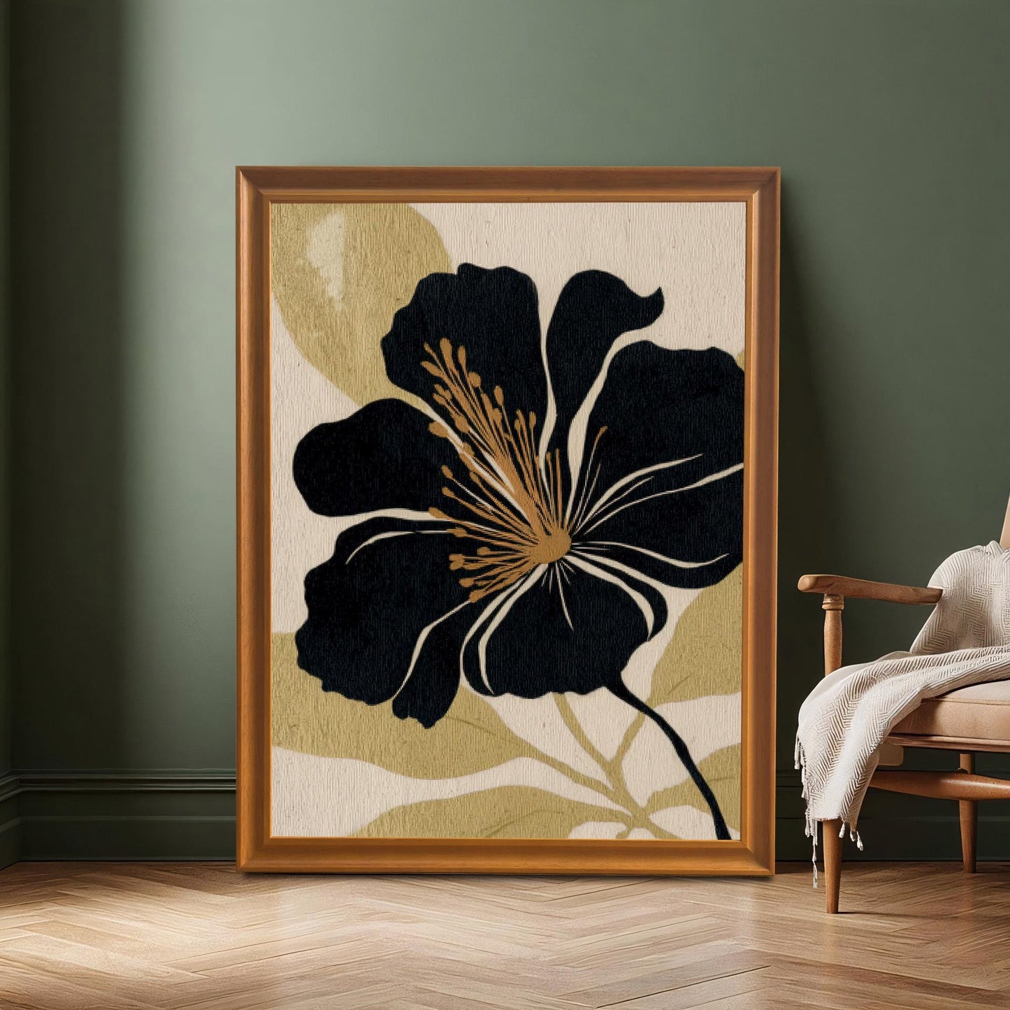 A large black and gold floral art print in a gold-colored frame resting on a wooden floor against a dark green wall, next to a vintage wooden armchair with a light-colored throw blanket.