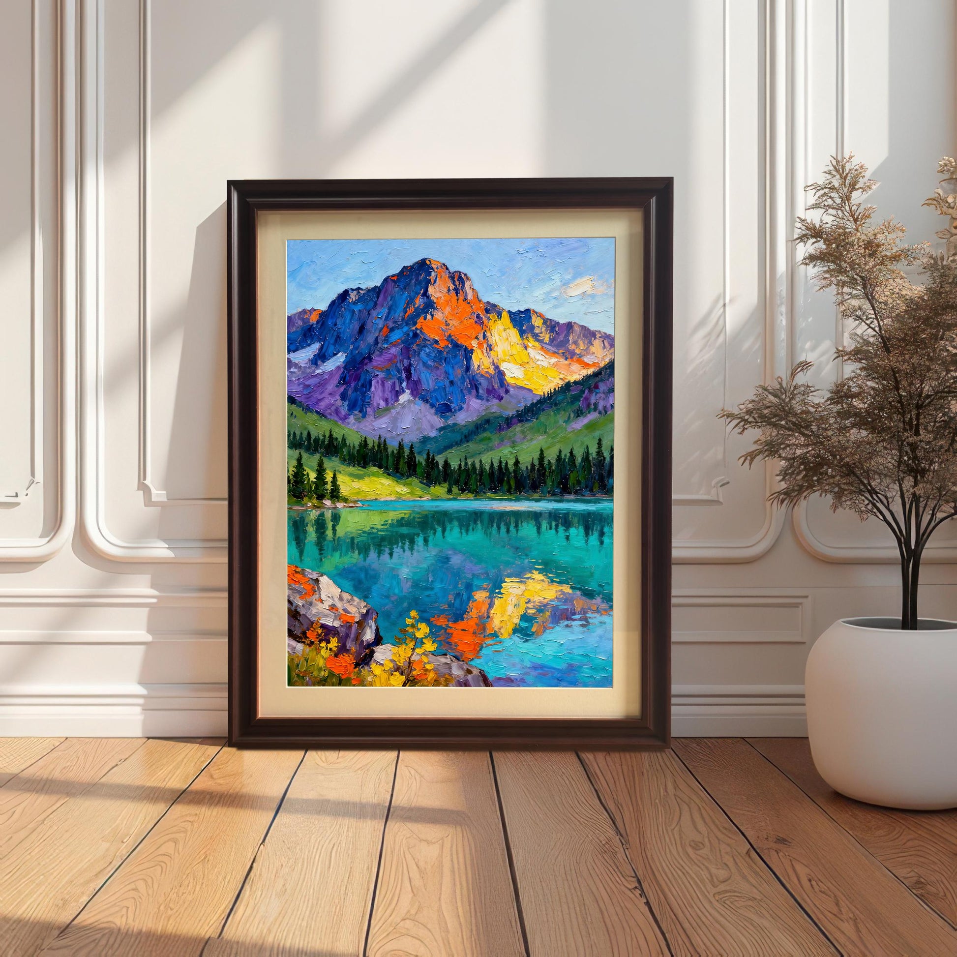 A vibrant oil painting print of a colorful mountain and lake landscape. The print is shown in a dark brown/coffee frame with a cream mount/mat, leaning against a white decorative wainscoting wall on a wooden floor.