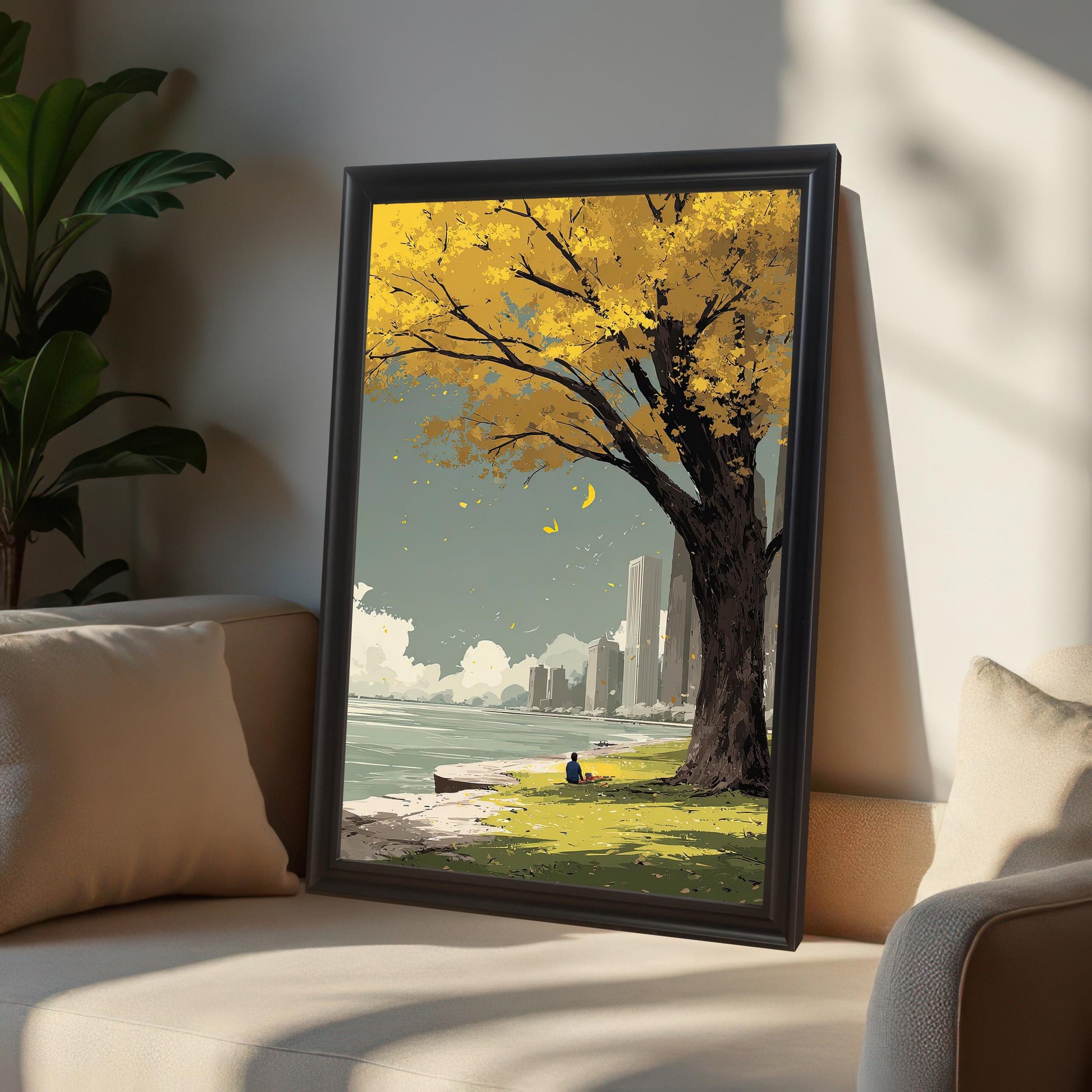 Framed art print of a yellow autumn tree and a city skyline reflected in water. The print is in a black wood frame and is leaning on a neutral-colored sofa with a large house plant in the background.
