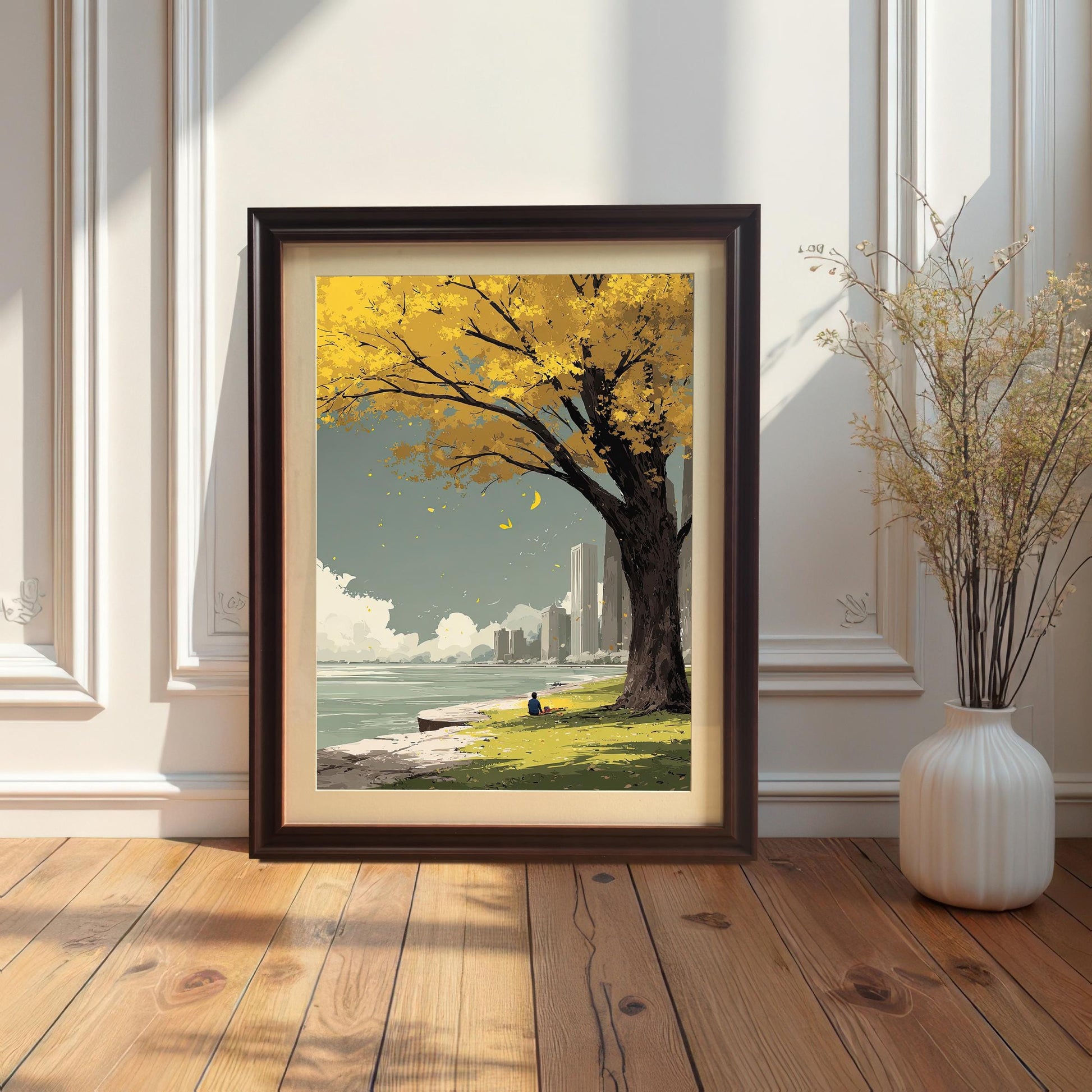 Framed art print of a yellow autumn tree and a person sitting by a lake with a cityscape. The print is in a dark brown/coffee frame with a cream mount/mat and is leaning against a white wainscoting wall on a wood floor next to a white vase.