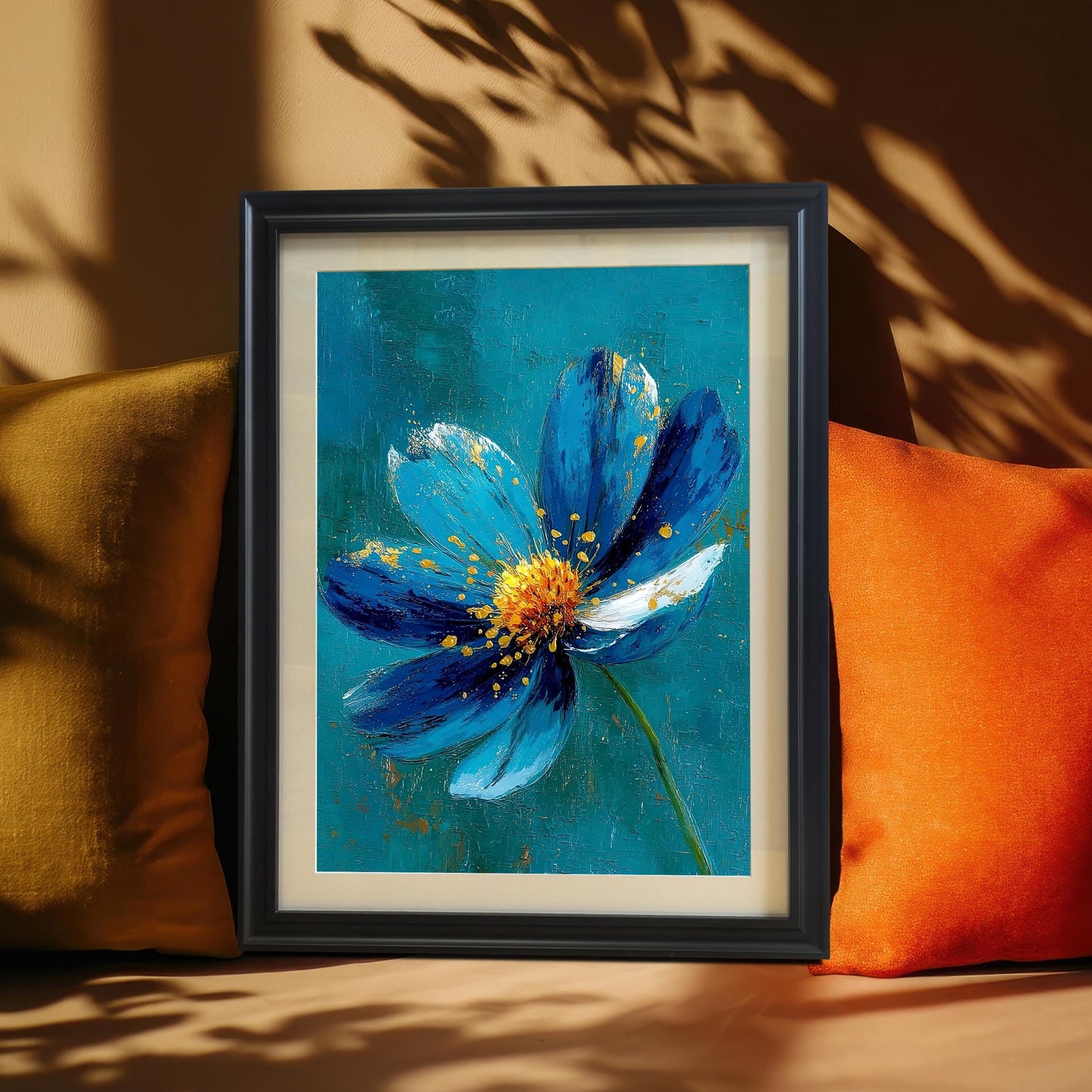A framed art print of an abstract blue and gold flower painting, set in a Black frame with a mount (mat), leaning between an orange and a yellow cushion. Cast shadows create a dramatic backdrop.