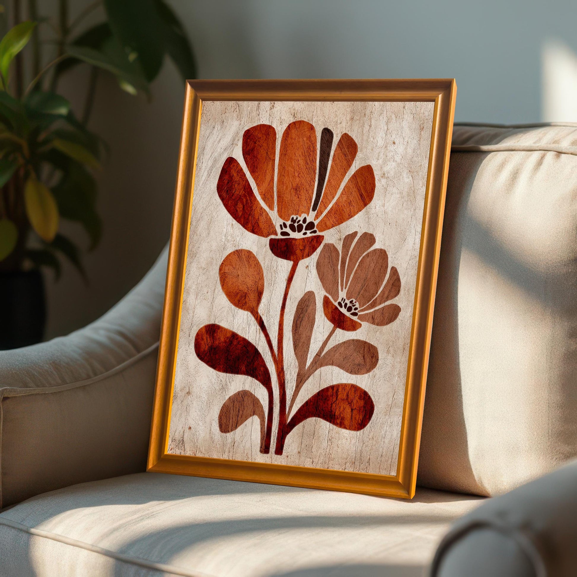 Framed rust-orange floral art print with an orange/gold frame resting on the arm of a beige upholstered chair, with sunlight streaming across it. (This is another lifestyle shot, showing the art in a home environment.)