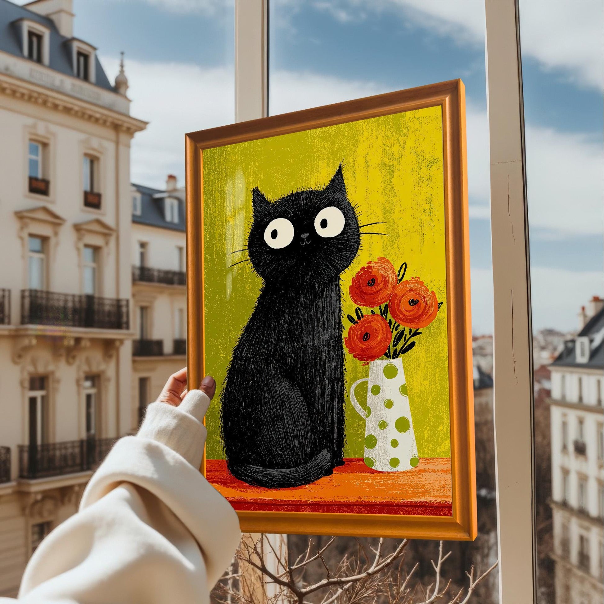 A hand holds the framed black cat art print with an Orange/Gold frame up to a sunny window overlooking European apartment buildings, showing the vibrant colors in natural light.