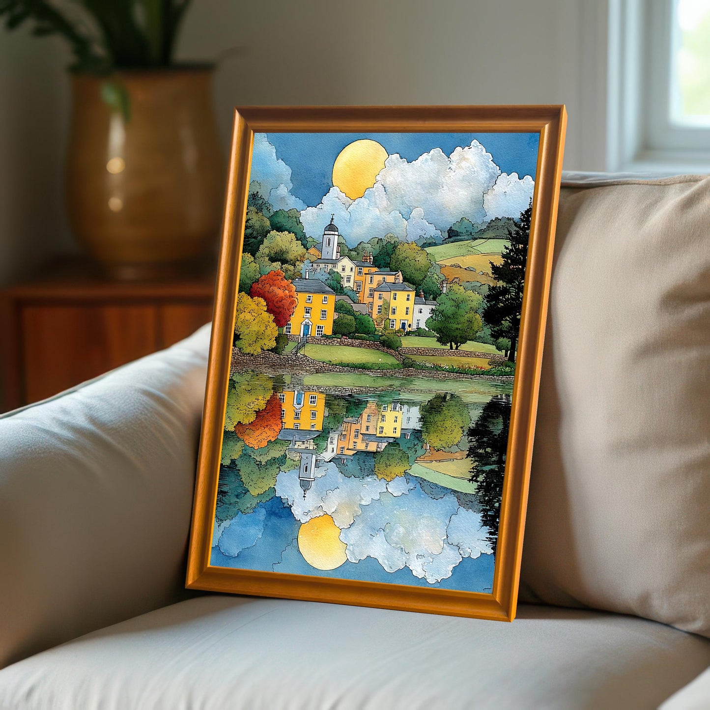 Framed art print propped on a light-colored couch next to a window. The frame is a warm orange or gold color. The scene is a reflective, colorful village landscape.