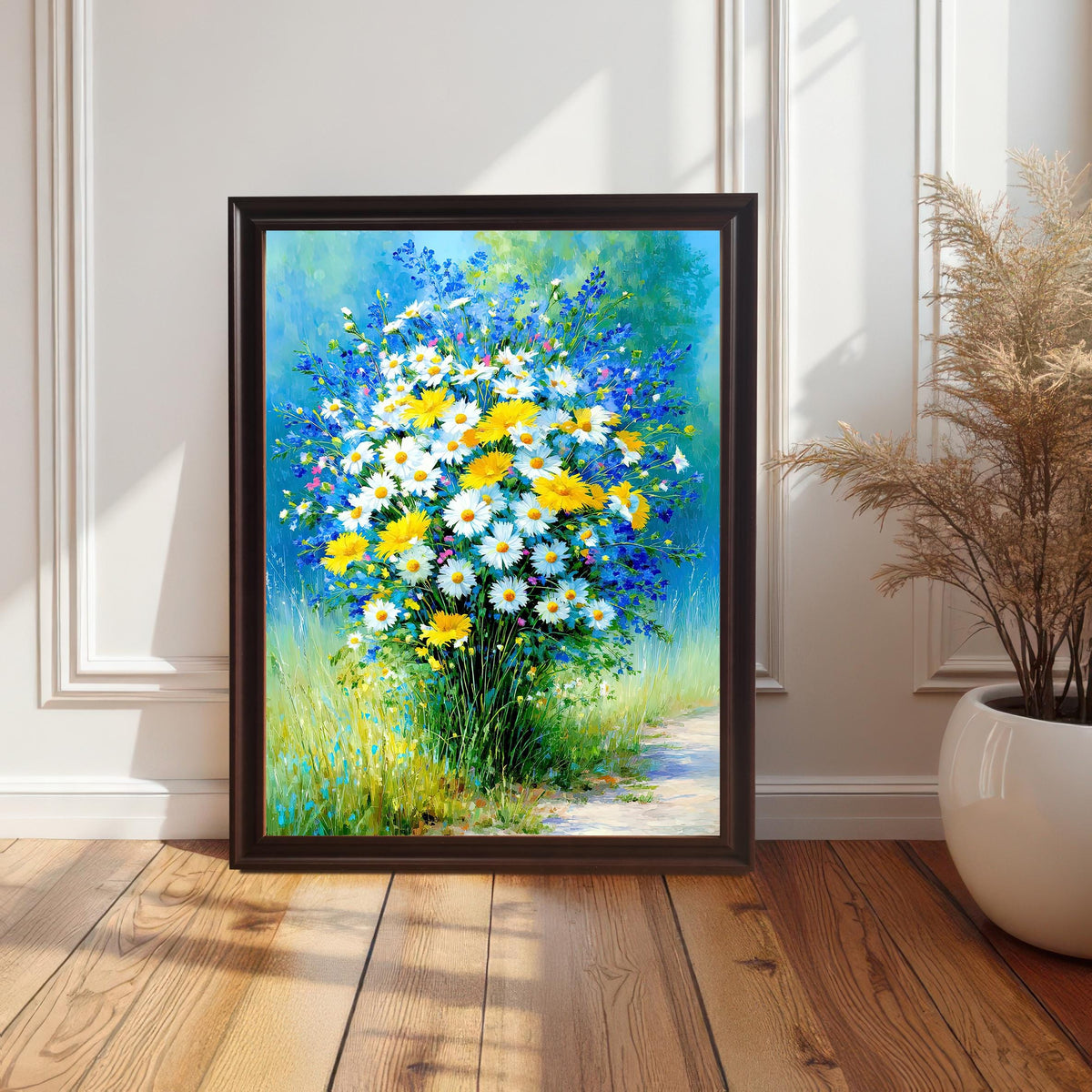 A vertically framed art print with a dark brown or coffee wood frame, leaning on a white wainscoted wall. The print is a vibrant impressionistic painting of a field bouquet with white and yellow daisies and blue flowers.