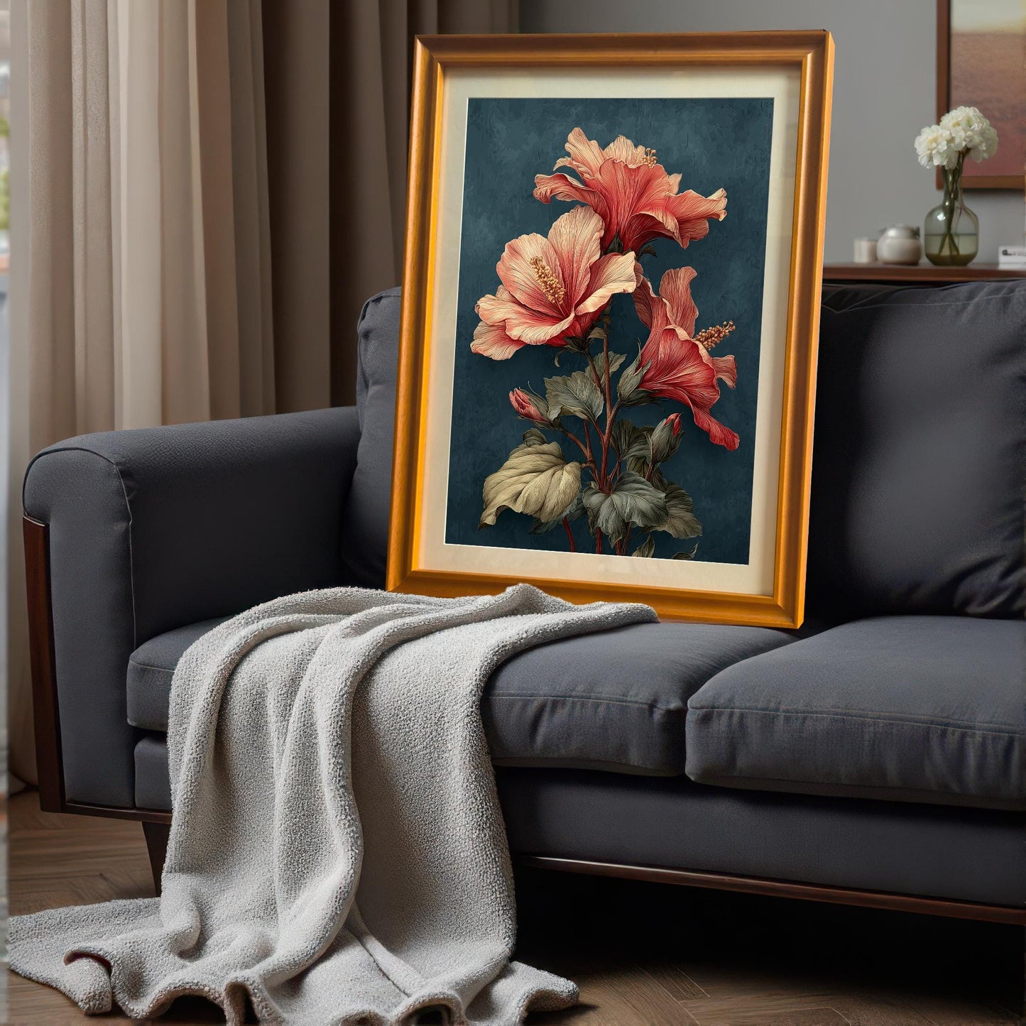 Framed art print with a gold or orange frame leaning on a dark grey sofa with a throw blanket. The art is a striking vertical print of coral hibiscus flowers on a deep teal background.