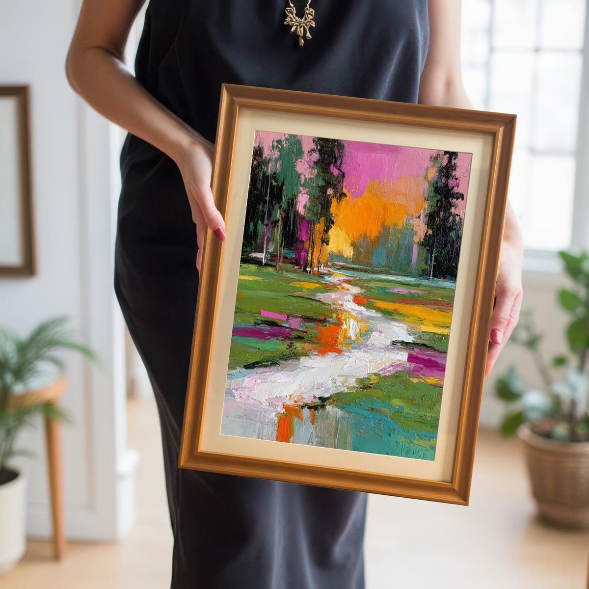 Woman in a black dress holding a small, gold-framed abstract landscape print with a stream, showing a close-up of the artwork and its gold frame