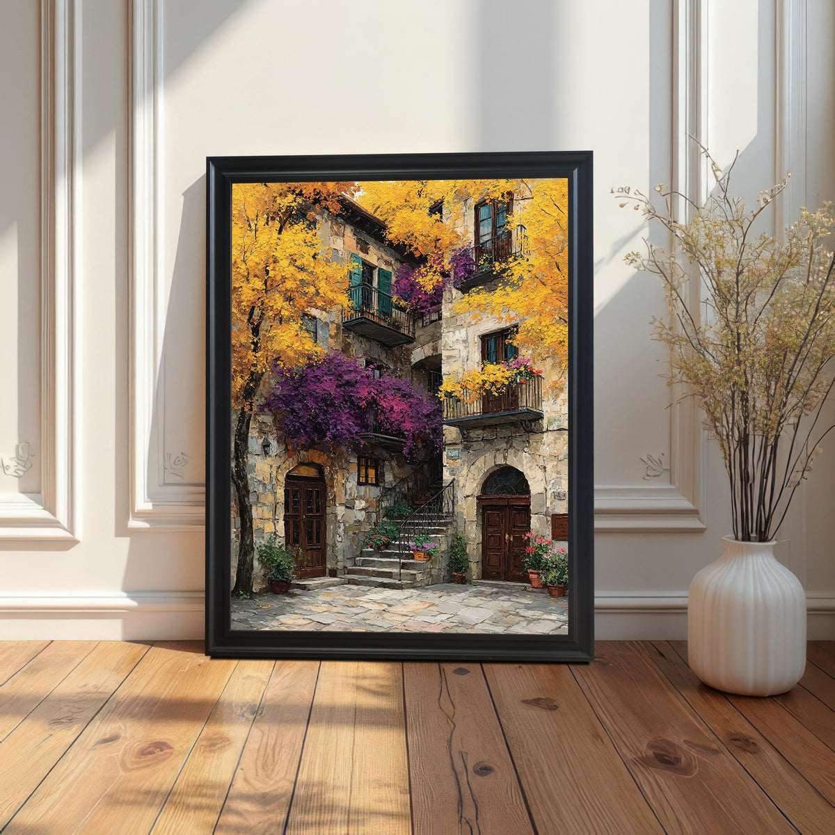 Framed art print with a black wood frame leaning on a light wooden floor against a white wainscoted wall next to a white vase with dried reeds.