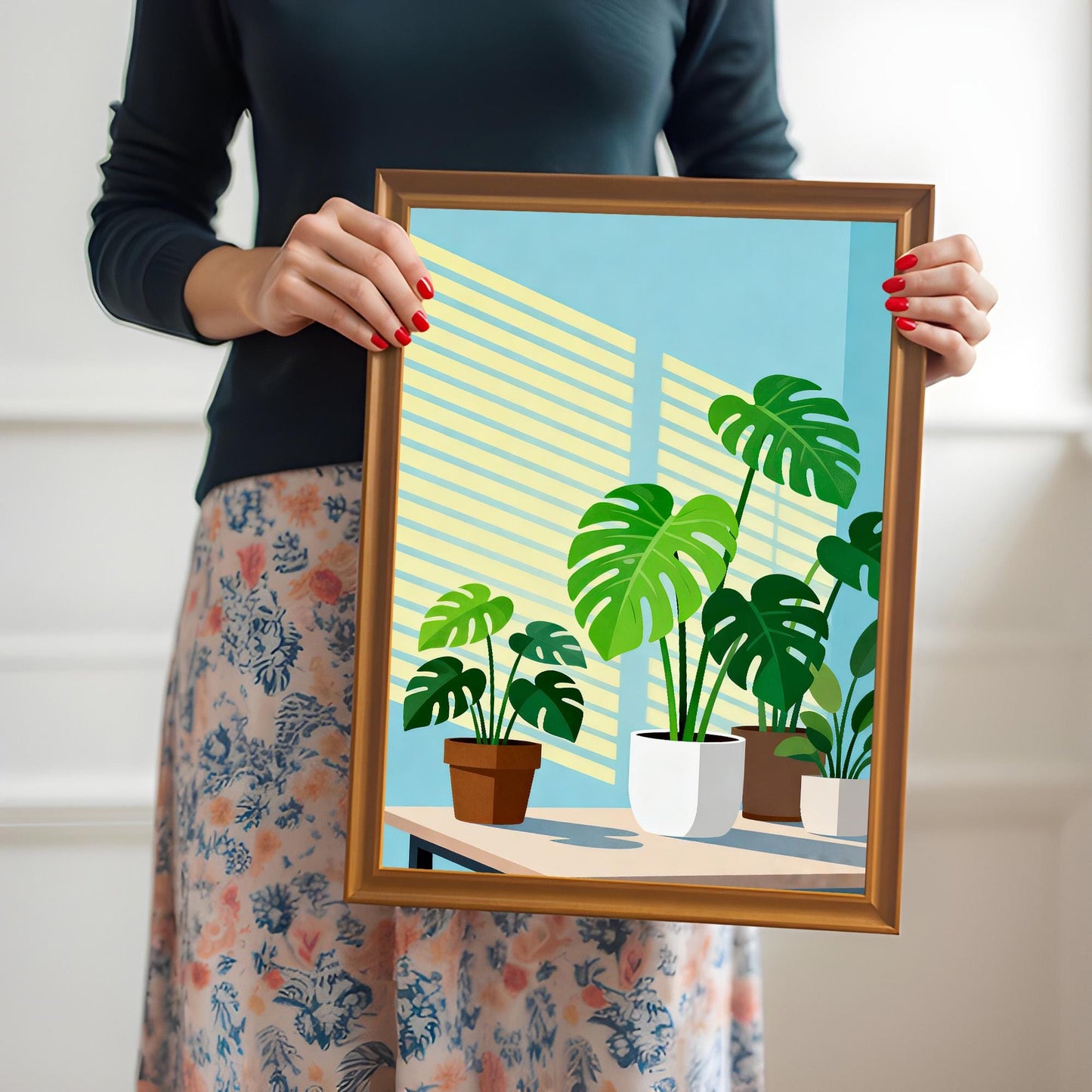 A person holding a framed 'Monstera Plants' art print in a brown Orange Frame. The person is wearing a dark top and a floral skirt and has red nail polish.
