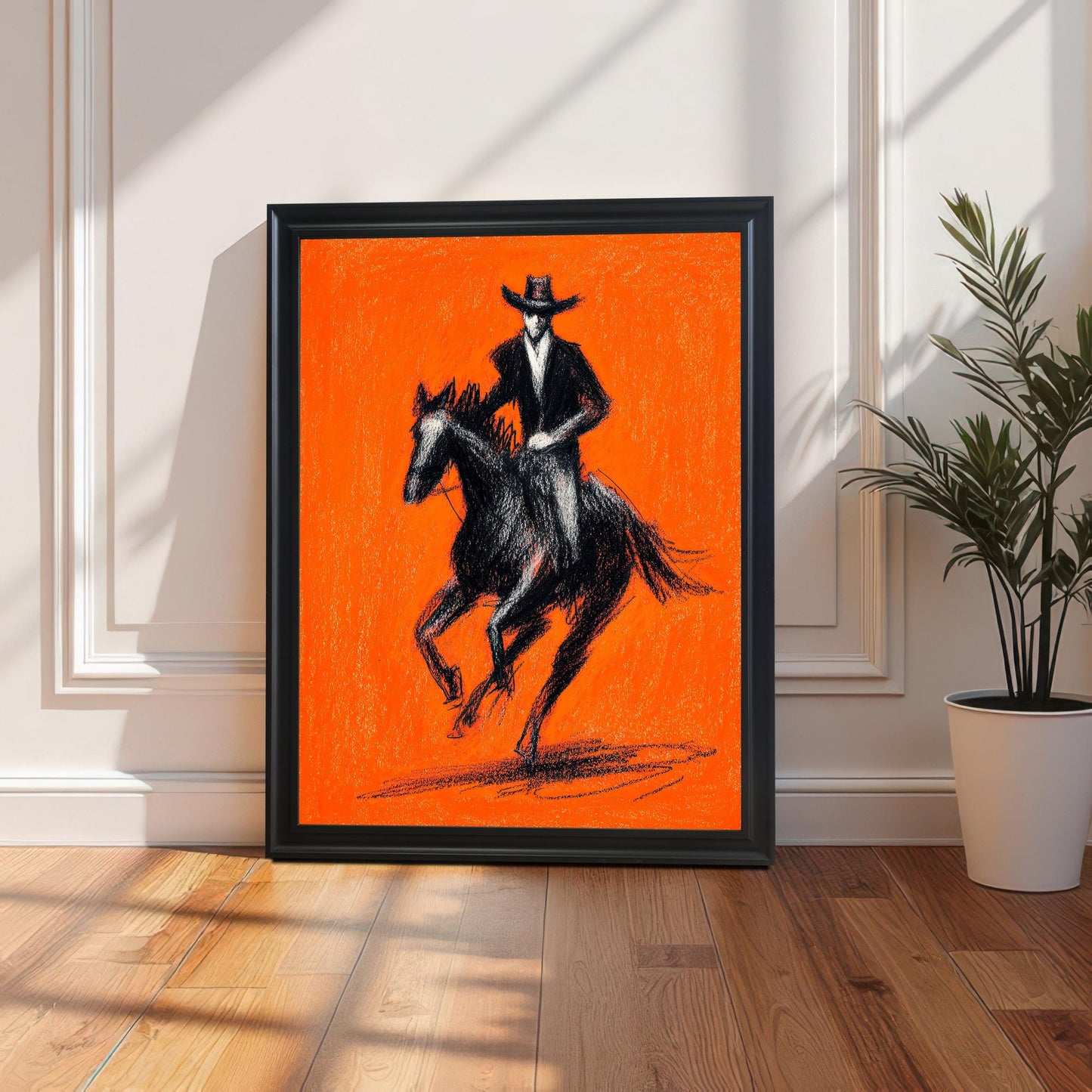 Lifestyle shot of the black and orange cowboy and horse art print with a Black wood frame, leaning on a light-colored wood floor against a white paneled wall. Sunlight casts a strong shadow across the floor. A tall, leafy potted plant sits to the right of the artwork.