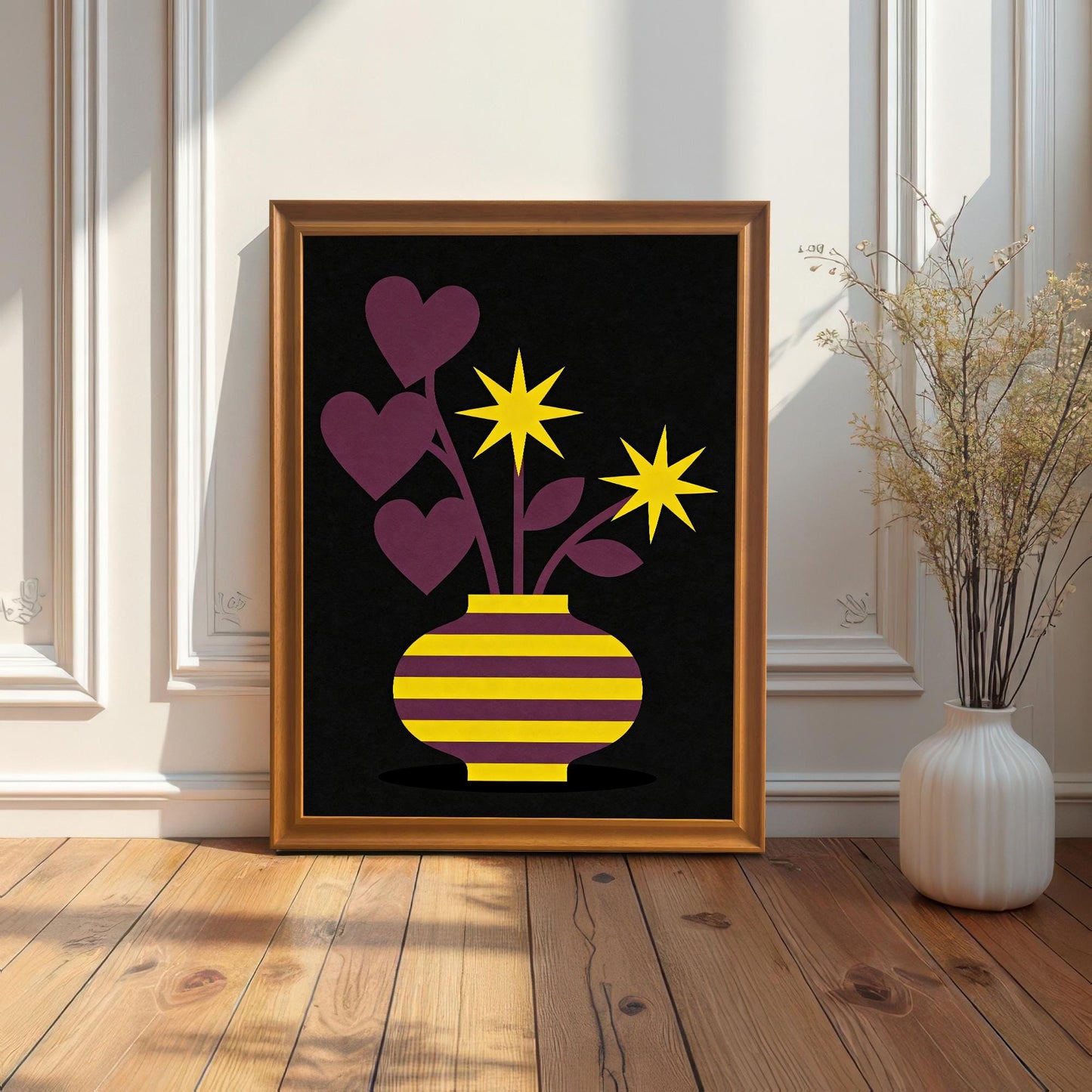 A gold-toned Orange framed art print featuring a purple and yellow striped vase with purple heart-shaped leaves and yellow star flowers on a black background. The framed print is resting on a wood floor against a white paneled wall next to a tall, white vase with dried reeds.