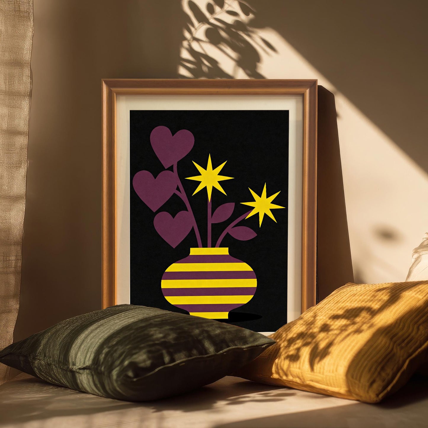 A gold-toned framed art print featuring a striped vase with heart-shaped leaves and star flowers, propped up on a surface among dark green and yellow pillows with sunbeams and shadows falling across it.