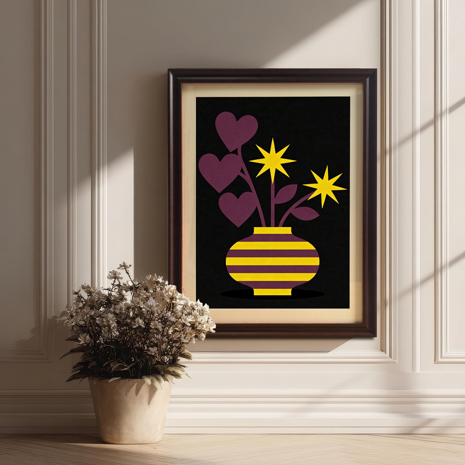 A dark brown Coffee framed art print featuring a purple and yellow striped vase with purple heart-shaped leaves and yellow star flowers on a black background. The framed print is resting on a wood floor against a white paneled wall next to a potted plant with dried white flowers.