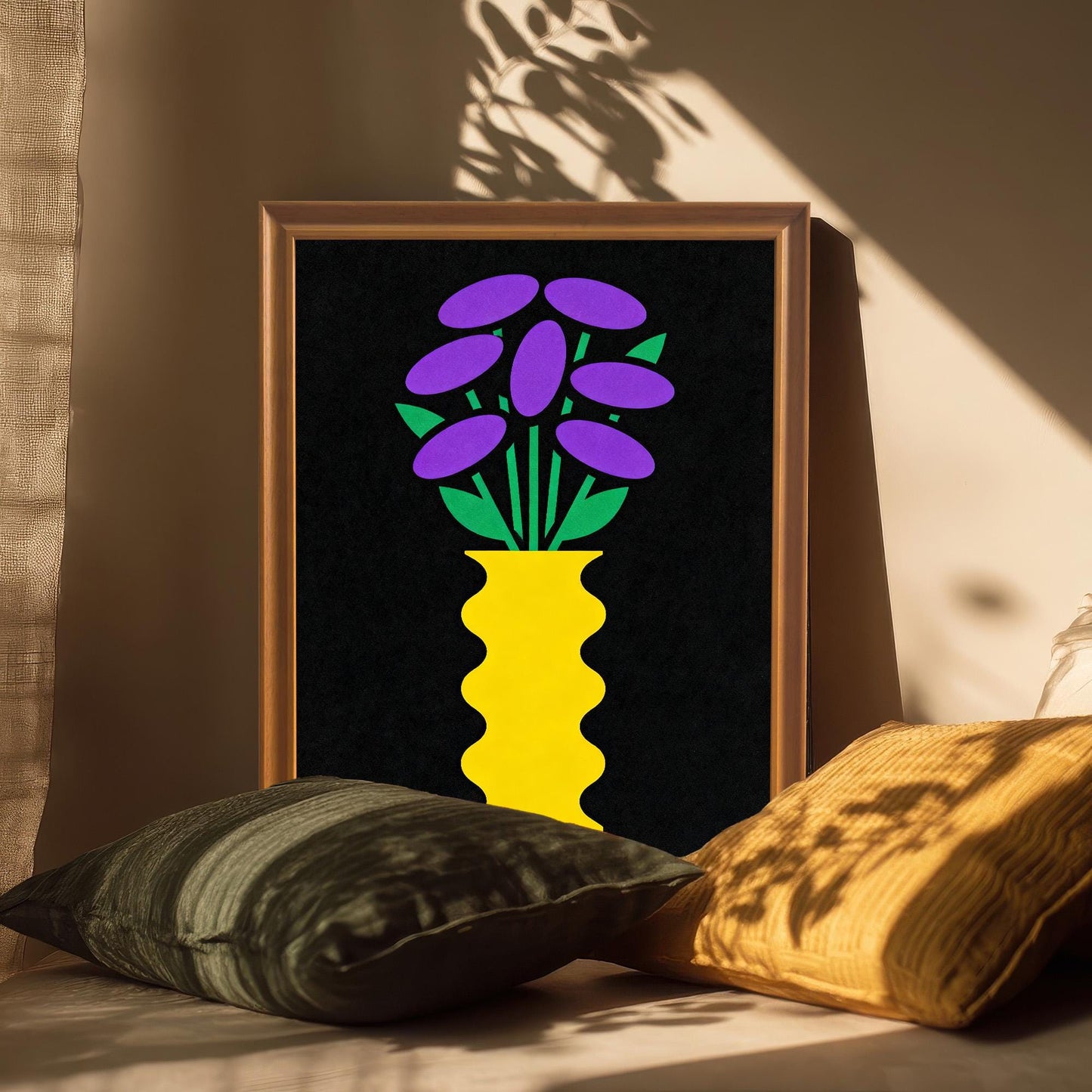 A gold-toned framed art print featuring a bright yellow vase with purple and green flowers on a black background. The print is angled and propped up on a surface among soft pillows, with sunbeams and shadows falling across it.