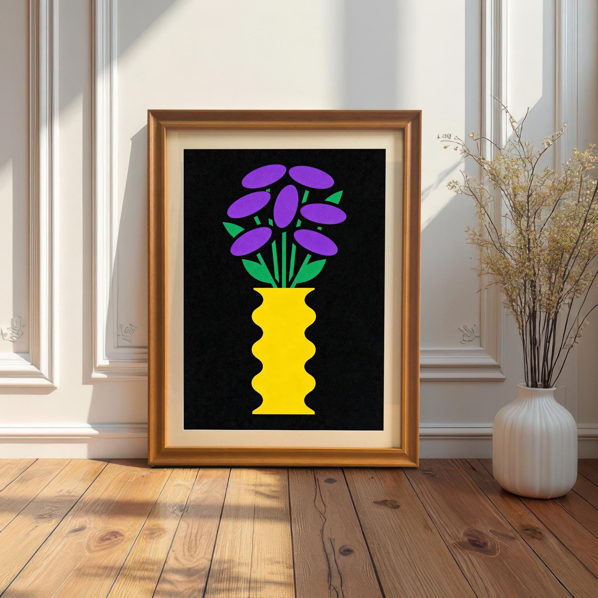 A gold-toned Orange framed art print featuring a bright yellow vase with purple and green flowers on a black background. The framed print is resting on a wood floor against a white paneled wall next to a tall, white vase with dried reeds.