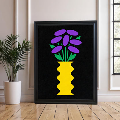 A large Black framed art print featuring a bright yellow, wavy vase with stylized purple and green flowers on a black background. The print is resting on a wood floor against a white paneled wall next to a tall houseplant in a white pot.