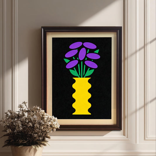 A coffee-brown framed art print featuring a bright yellow vase with purple and green stylized flowers on a black background. The framed print is resting on a light wood floor against a white paneled wall, with a potted plant with dried white flowers next to it.
