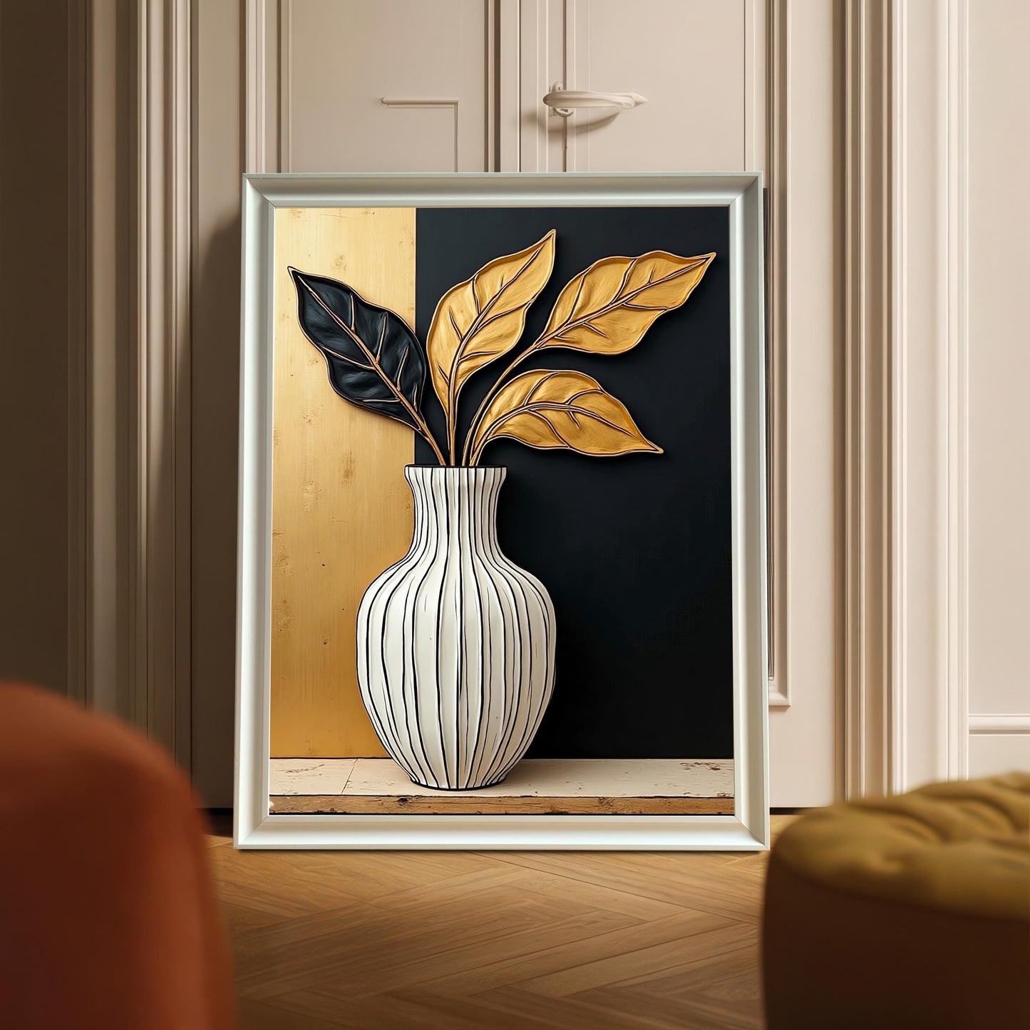 White framed art print featuring a white striped vase with black and gold leaves, set against a split gold and black background. The print is leaning on a wooden floor in front of light-colored cabinets, with a yellow tufted footrest visible.
