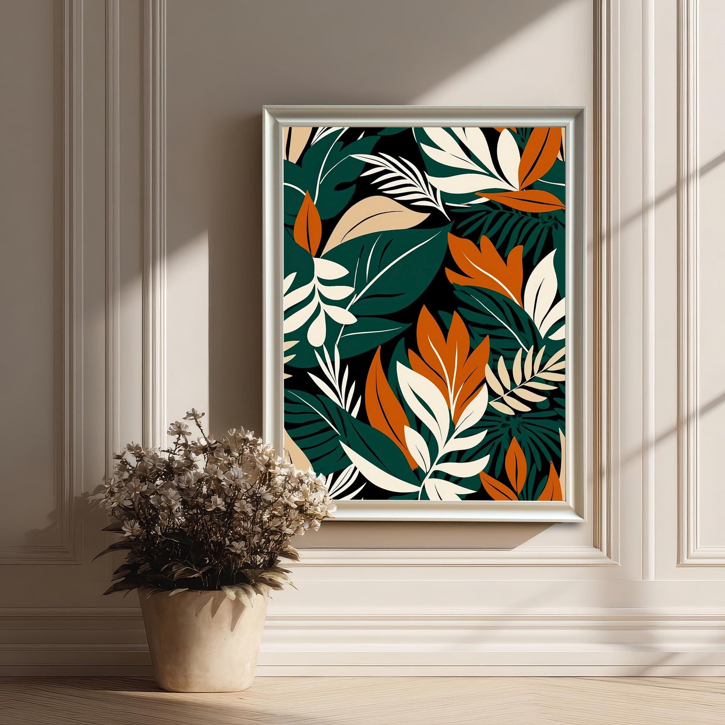 Styled image of the tropical leaf art print framed in White on a panelled, off-white wall. Sunlight casts shadows, and a dried floral arrangement in a pot is in the foreground.