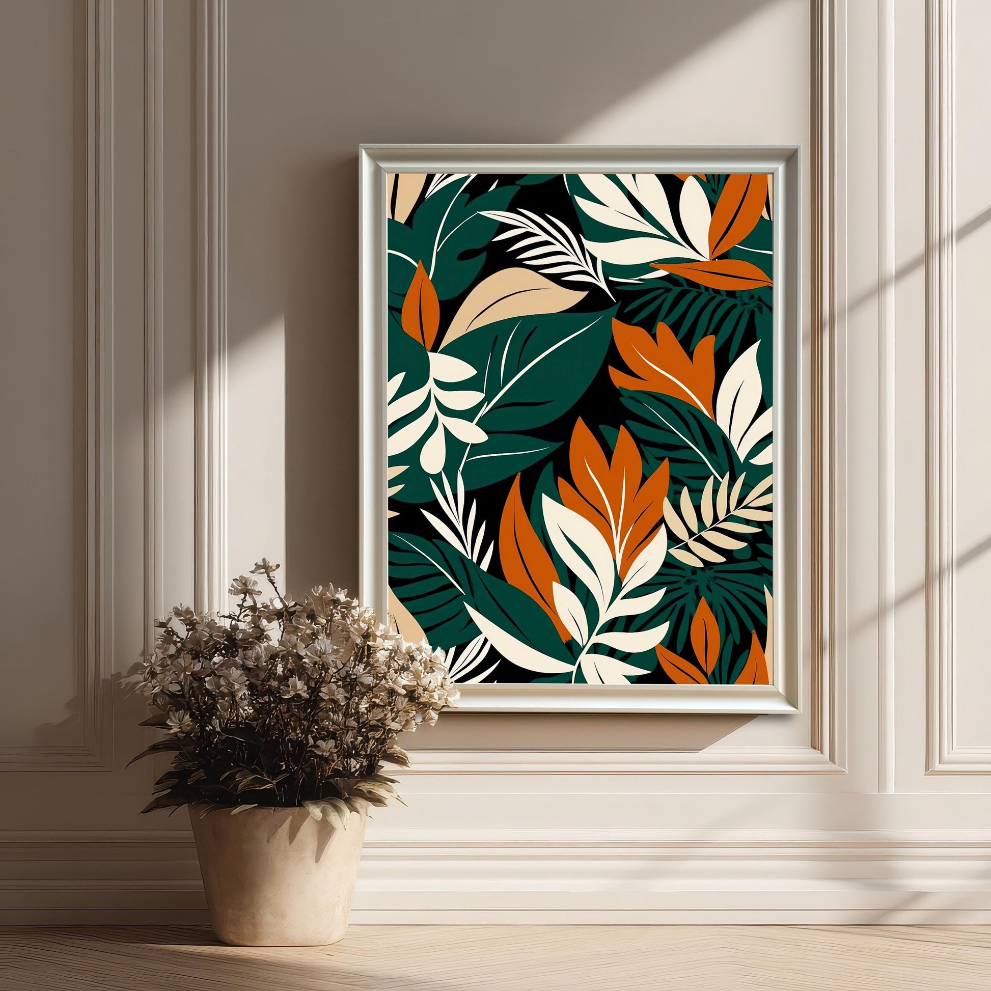 Styled image of the tropical leaf art print framed in White on a panelled, off-white wall. Sunlight casts shadows, and a dried floral arrangement in a pot is in the foreground.