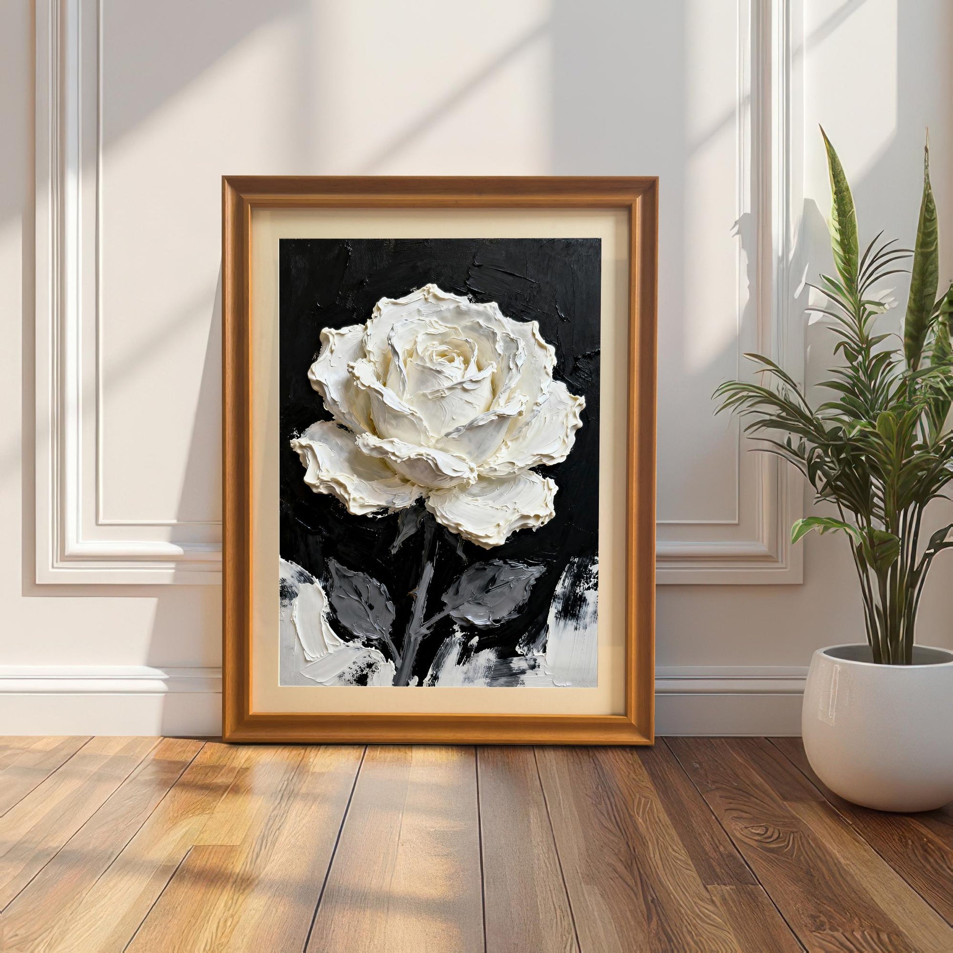 A framed print of the textured white rose painting with an Orange wood frame and a mount (matting), leaning on a wooden floor against a white wall with ornate molding. A potted snake plant is to the right.
