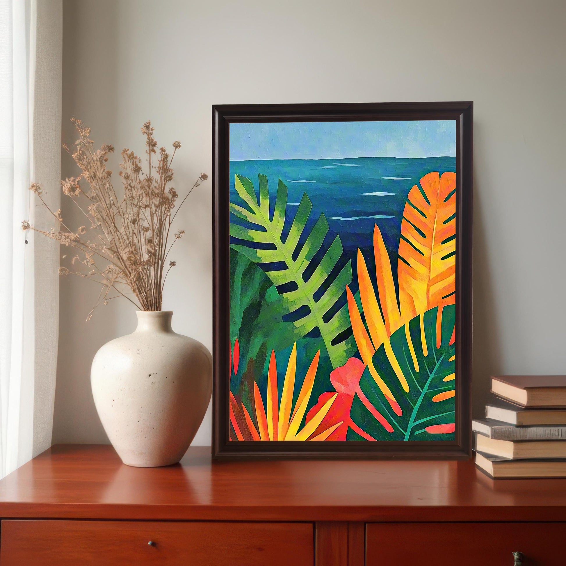 A lifestyle image of the framed art print of the tropical leaves overlooking the blue ocean. The print is in a Coffee Wood (dark brown) frame with a mount and is displayed on a dark wood dresser next to a cream vase of dried flowers and a stack of books.