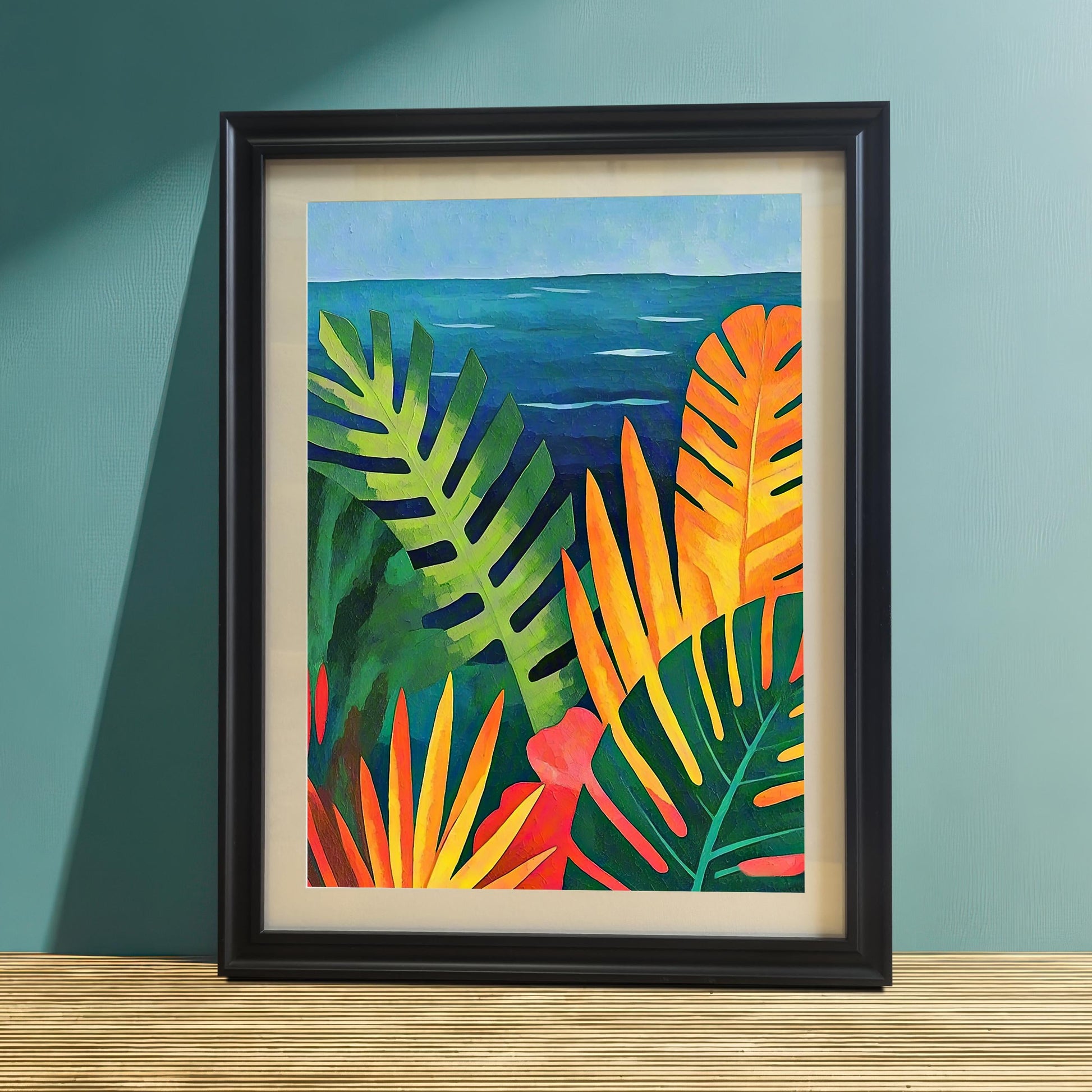 A close-up studio shot of the vibrant tropical foliage and ocean art print in a Black Wood Frame, leaning on a light wood surface against a teal-colored wall. Sunlight and shadow highlight the colorful scene.