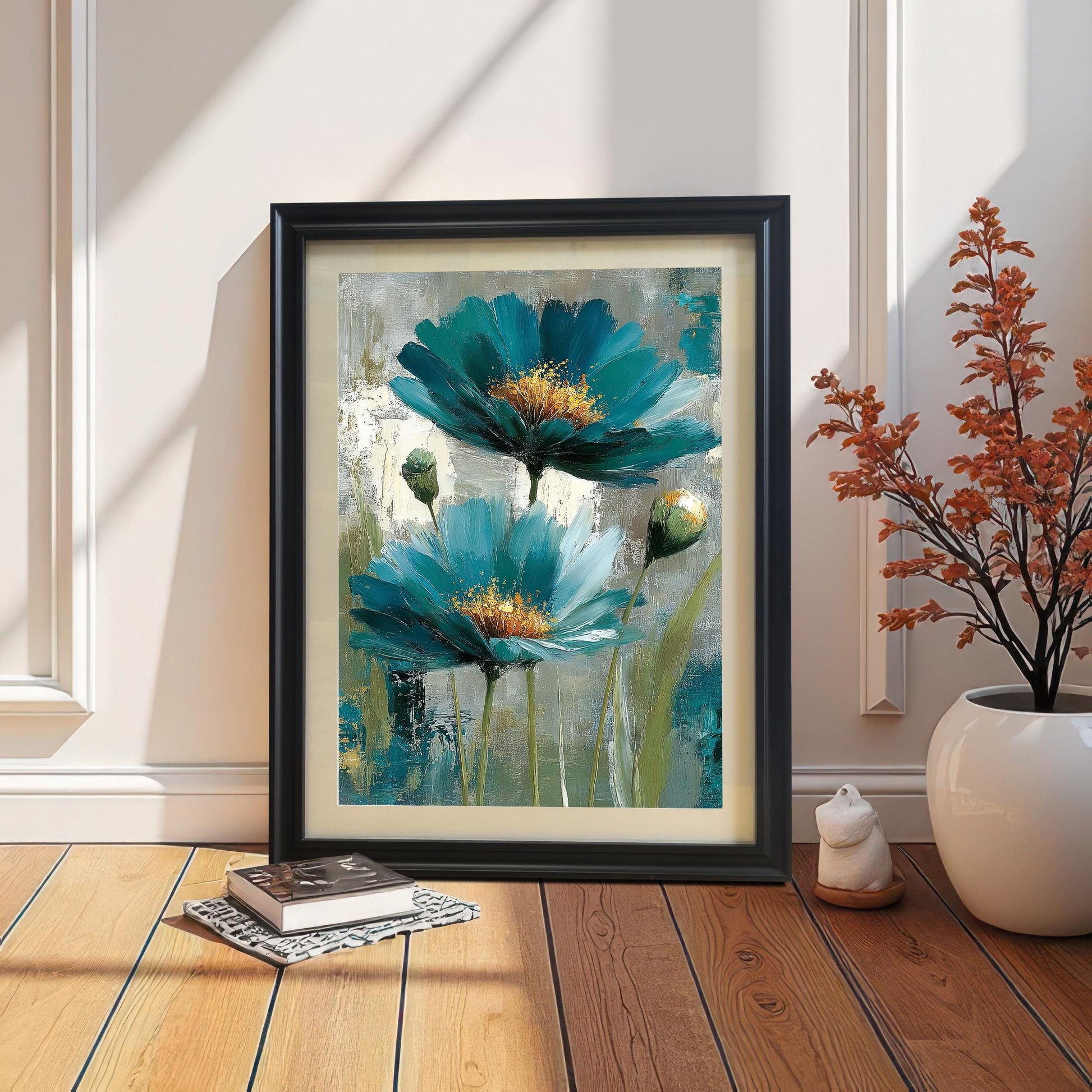 A framed art print in a Black wood frame with a mount leaning on a wooden floor against a wainscoted wall. The print, featuring a painting of two abstract teal flowers, is next to a book and a potted plant with dried orange branches.