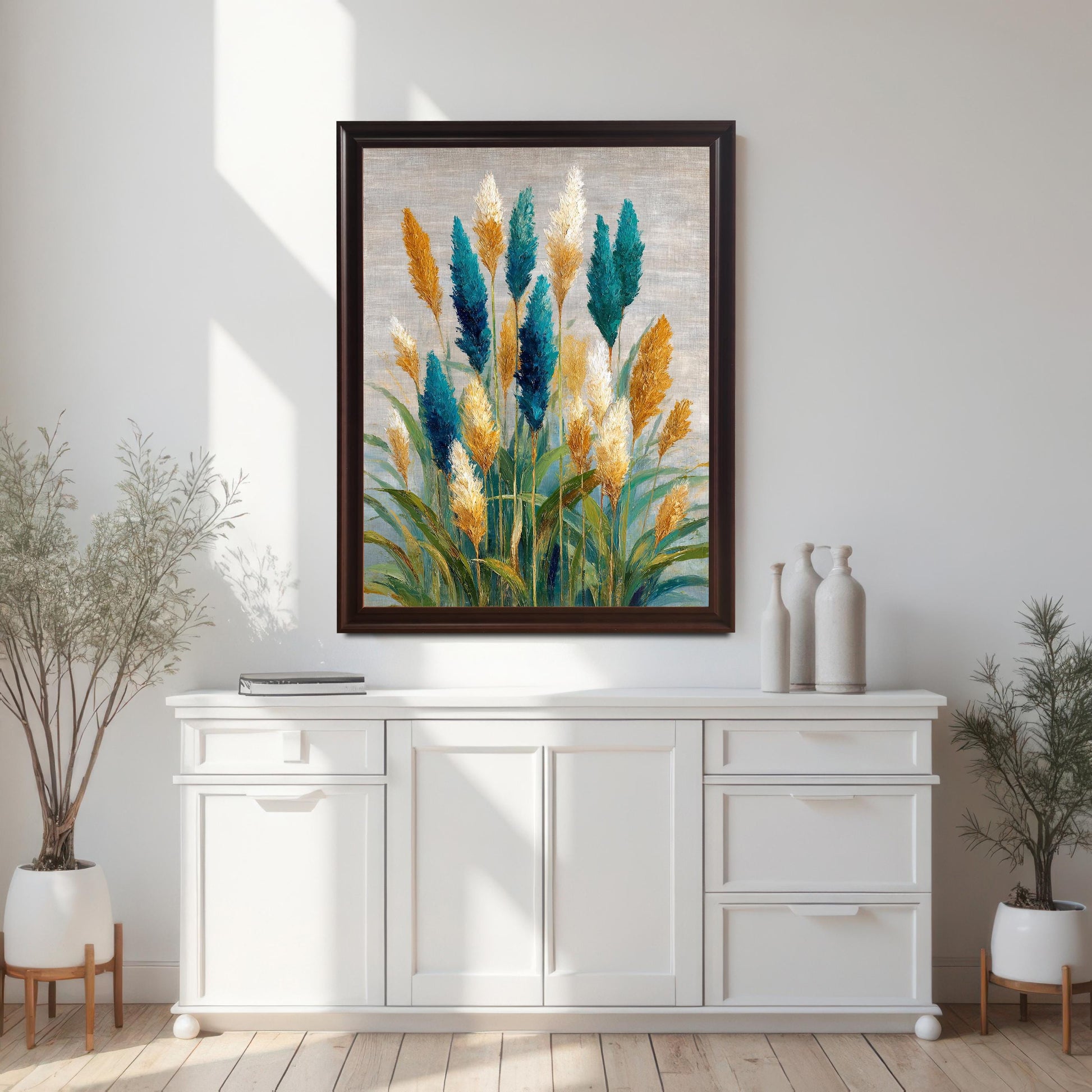 Wood-framed art print featuring vibrant, stylized pampas grass in teal, golden yellow, and white on a textured, neutral background. The artwork is hung above a white wood buffet or console table in a bright room with natural light and potted plants on both sides.