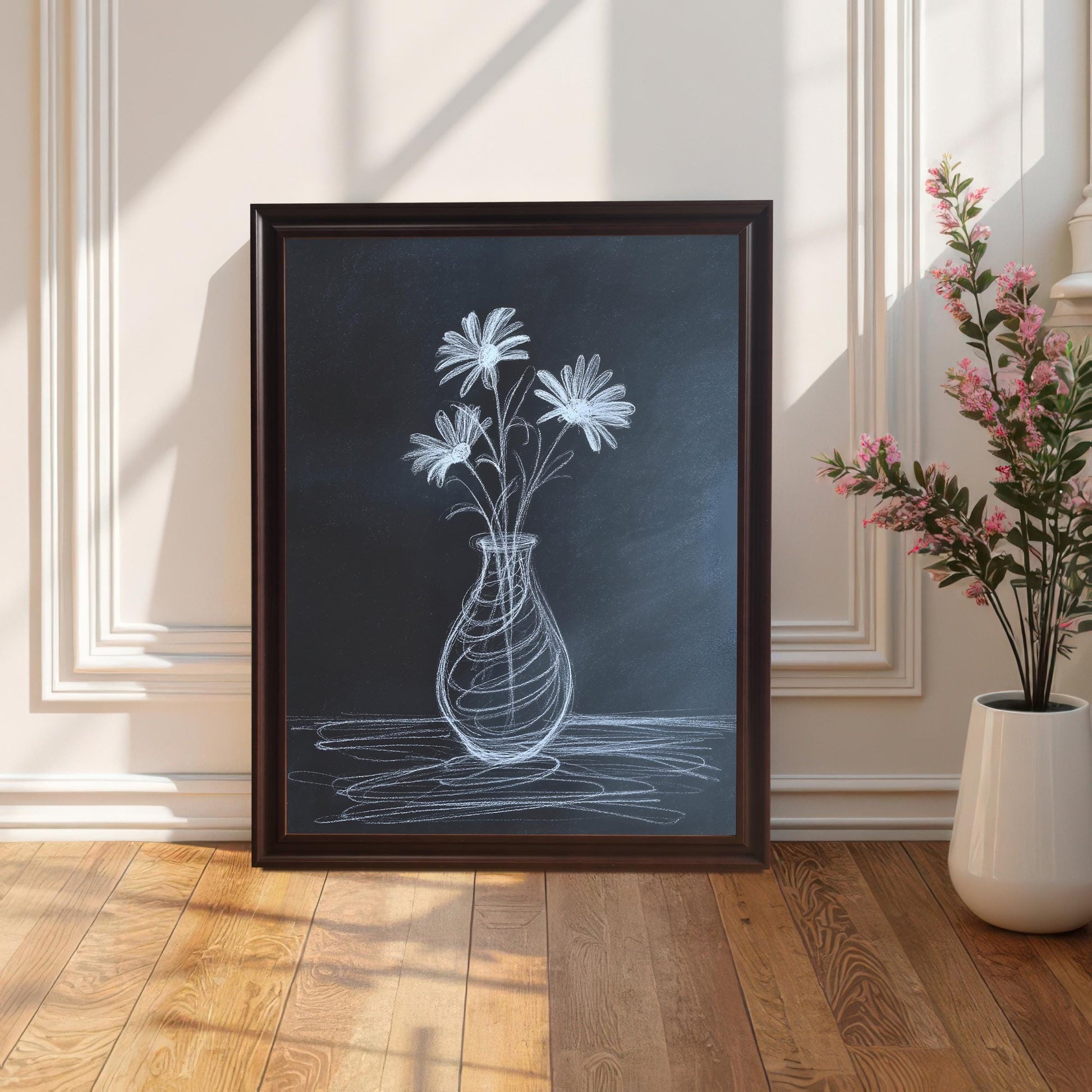 Full shot of the chalk flower print in a dark brown/coffee frame, leaning on a wooden floor against a white wall with wainscoting details, next to a tall vase of pink flowers.