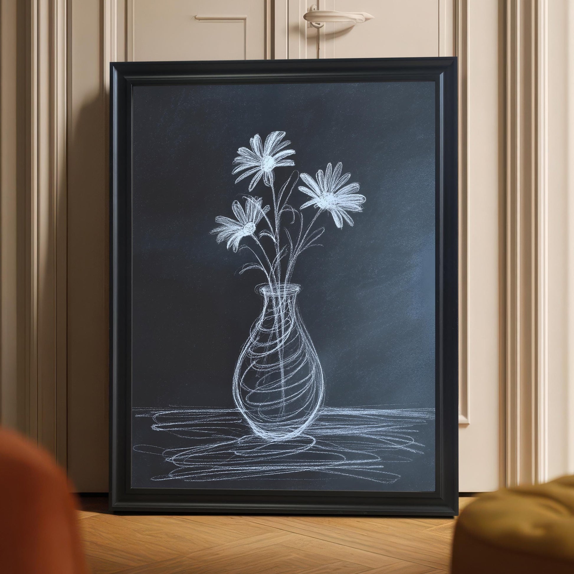Framed chalk flower print in a black wooden frame leaning on a light hardwood floor against a beige door, with the focus on the artwork. The scene includes hints of orange and yellow accent furniture in the foreground.