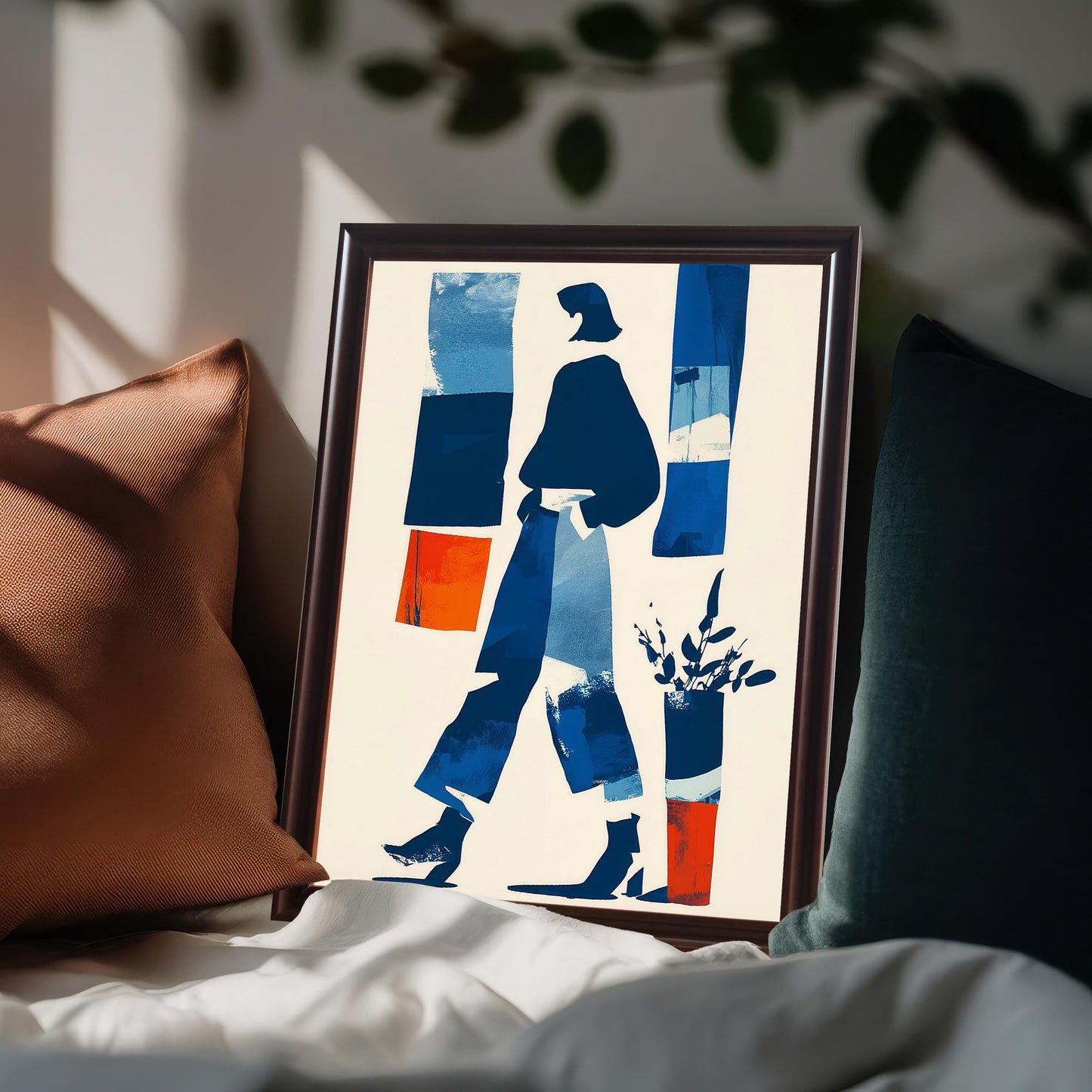 Close-up shot of a framed abstract print featuring a walking female figure in blue and white, with vertical blocks of dark blue, light blue, and orange. The dark brown framed art leans against pillows on a couch or bed.